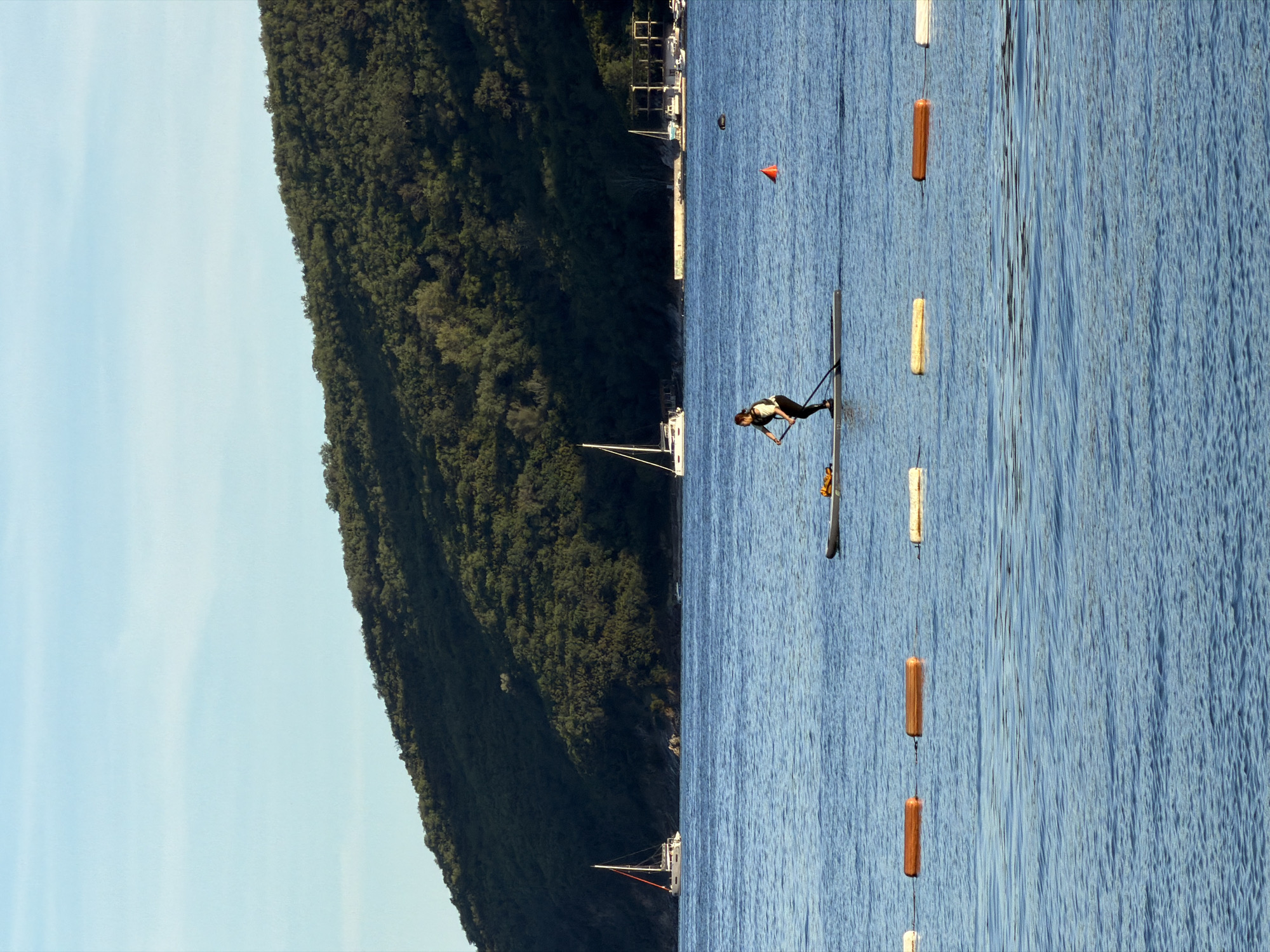 A kayaker paddles on the Adriatic Sea near Budva, Montenegro. The scene includes the coastline, densely covered with trees, meeting a blue sky on the left side. The sea occupies the right side of the image, with a person positioned towards the center of the frame on a narrow kayak. The kayak is situated between a series of floating buoys demarcating a lane. A sailboat and additional infrastructure are visible on the shoreline in the background.