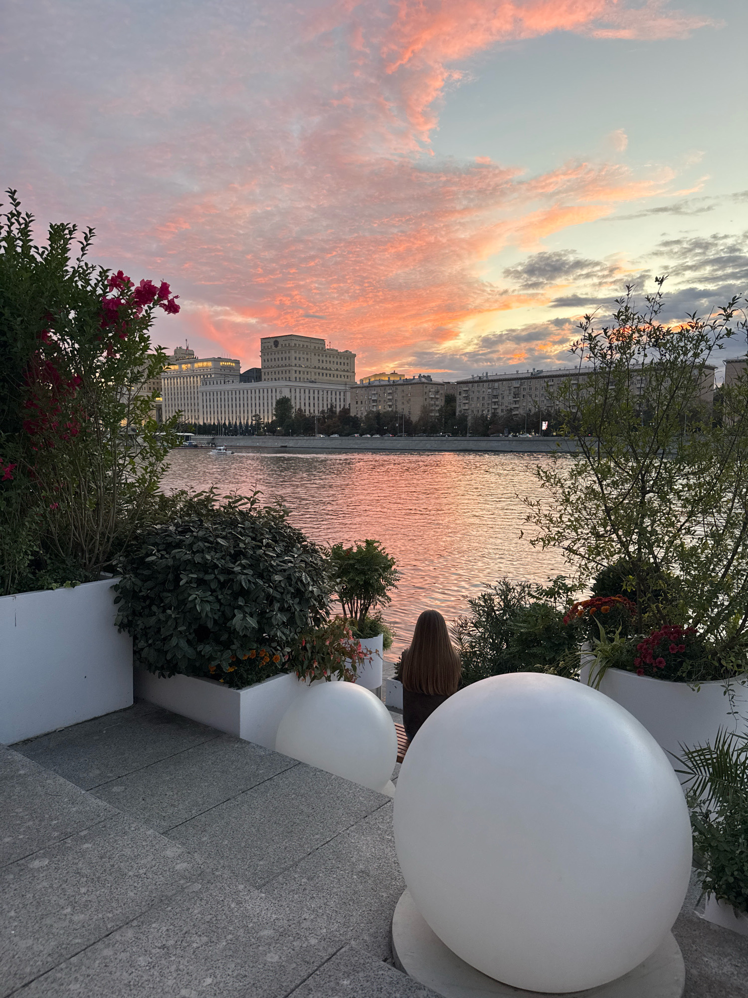 The image depicts a tranquil riverside scene at dusk in Moscow, Russia, likely on the Moskva River embankment. The sky is ablaze with a soft pink and orange sunset, with wispy clouds creating a dramatic backdrop. In the mid-ground, a wide river reflects the warm hues of the sky. Across the river, a grand, light-colored building, characteristic of Stalinist architecture, dominates the skyline, with smaller residential buildings flanking it.
In the foreground, a person with long brown hair is seated, facing away from the camera and towards the river. They are positioned behind lush greenery and white planters filled with flowers. The scene is framed by two large, white spherical objects, possibly decorative lights, and a granite staircase leading down. The overall atmosphere is serene and picturesque, capturing a moment of quiet contemplation by the water.