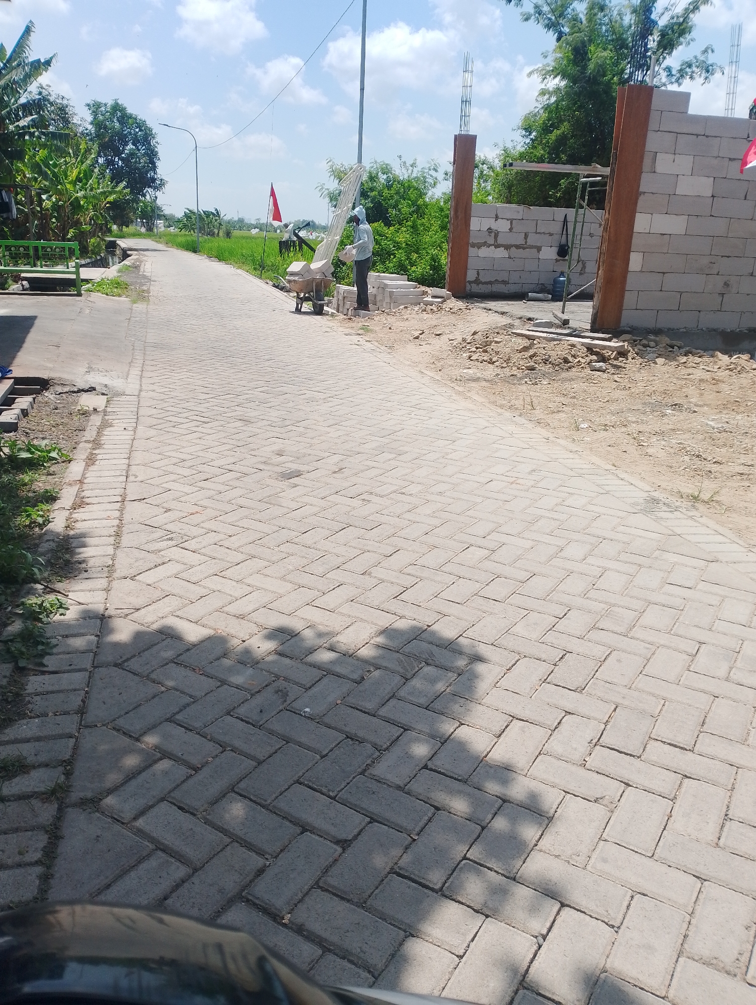 The image shows a paved road with interlocking bricks, typical of Indonesian street construction. On the right side of the road, a construction site is visible, with stacks of grey concrete blocks and wooden poles. A worker in a light blue shirt and white head covering is seen pushing a wheelbarrow filled with materials near a scaffolding. A red and white flag, likely the Indonesian flag, is planted near the construction site. To the left of the road, a green metal bench and some banana trees are present, bordering a patch of green rice paddies. The sky is blue with scattered white clouds. The overall scene suggests ongoing construction and development in a semi-rural area, possibly in Jumputrejo, Indonesia, given the context. The image captures a moment of daily activity and progress in a tropical environment.