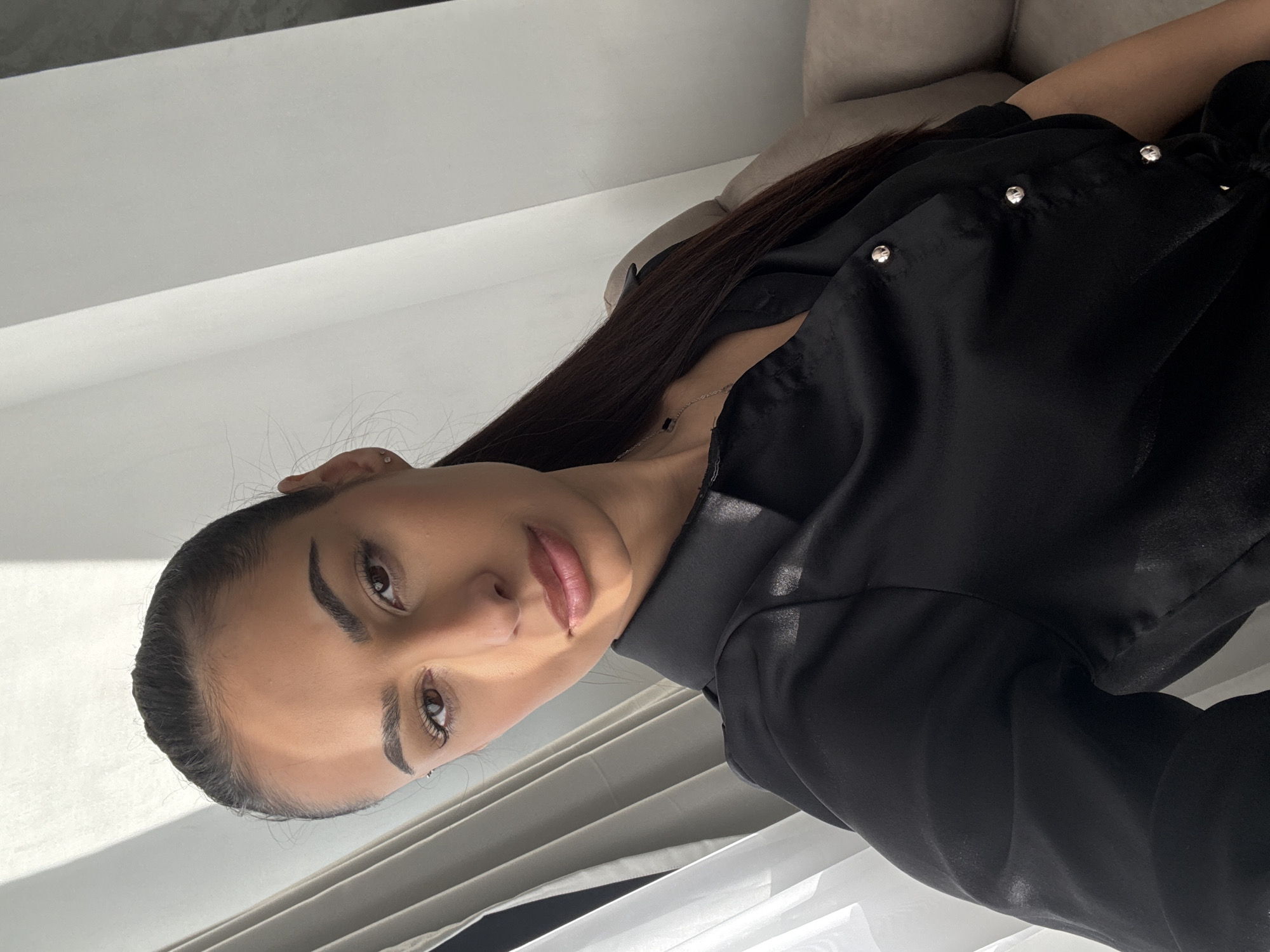 A young woman is posing for a selfie indoors. She has dark hair, tan skin, and is wearing a black, collared shirt. The background appears to be a wall and a couch. The location cannot be determined to be in Sofia, Bulgaria from the image alone.