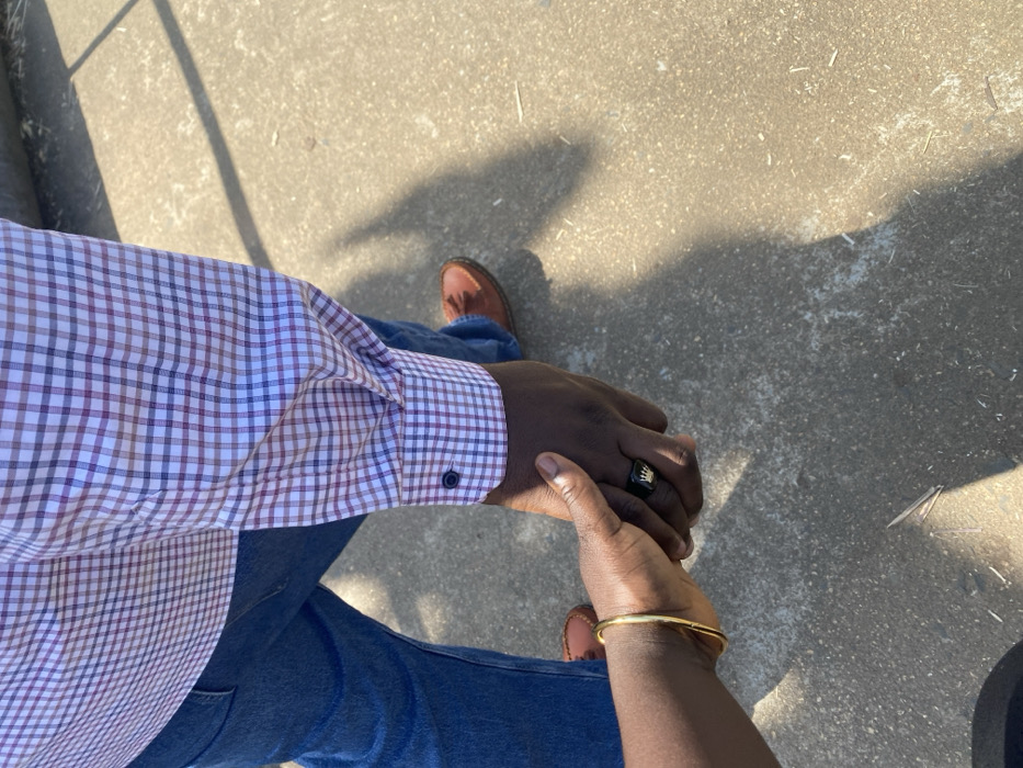 The image captures a close-up view of two individuals holding hands, set in Kpak, Nigeria. One person is observed from the mid-torso down, wearing a white, blue, and red plaid long-sleeve shirt, blue denim jeans, and brown leather loafer-style shoes. Their hand, adorned with a black ring featuring a gold crown emblem on the ring finger, is interlocked with the hand of a second person. This second individual's forearm and hand are visible, wearing a single gold-colored bangle on the wrist. Both individuals exhibit dark skin tones. They are standing outdoors on a light-colored, possibly concrete or paved, surface, under bright overhead illumination casting distinct shadows.