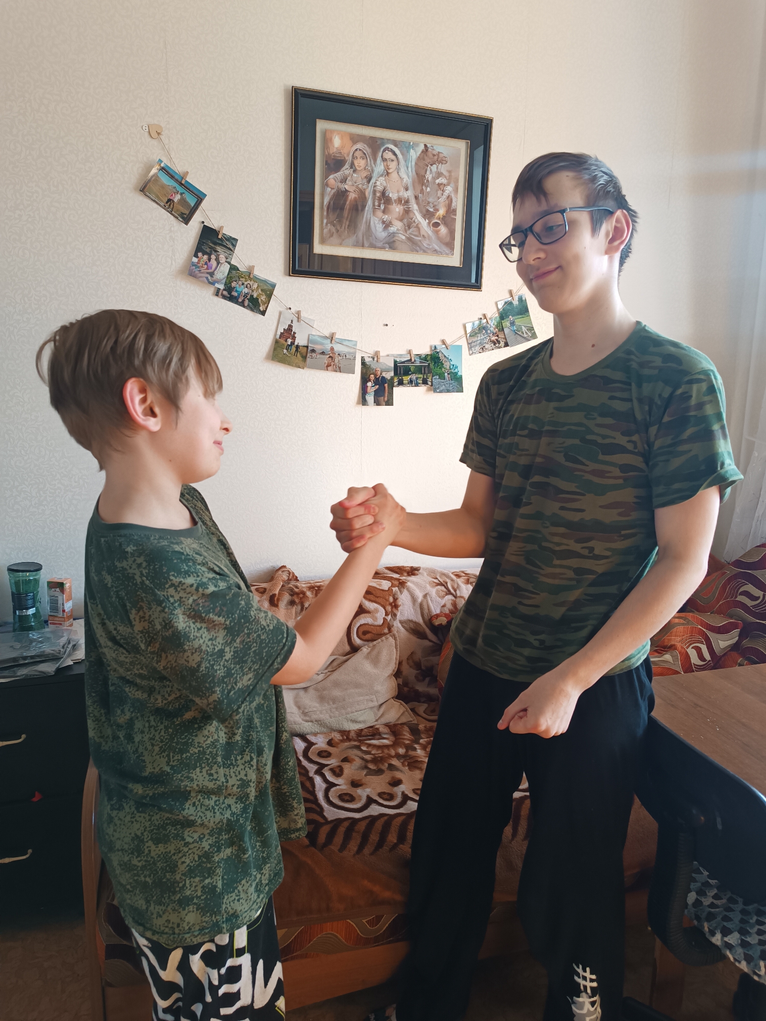 Two boys are standing indoors, engaged in a fist bump. The boy on the left has short brown hair and wears a green patterned T-shirt and patterned pants. The boy on the right is taller, wearing glasses, a camouflage T-shirt, and black pants.
Behind them is a wall decorated with a framed picture and a string of photographs attached with clothespins. A partially visible brown patterned couch is behind the boys. A cabinet with items on it is seen to the left, and a table is to the right. The location is Unknown, Unknown.