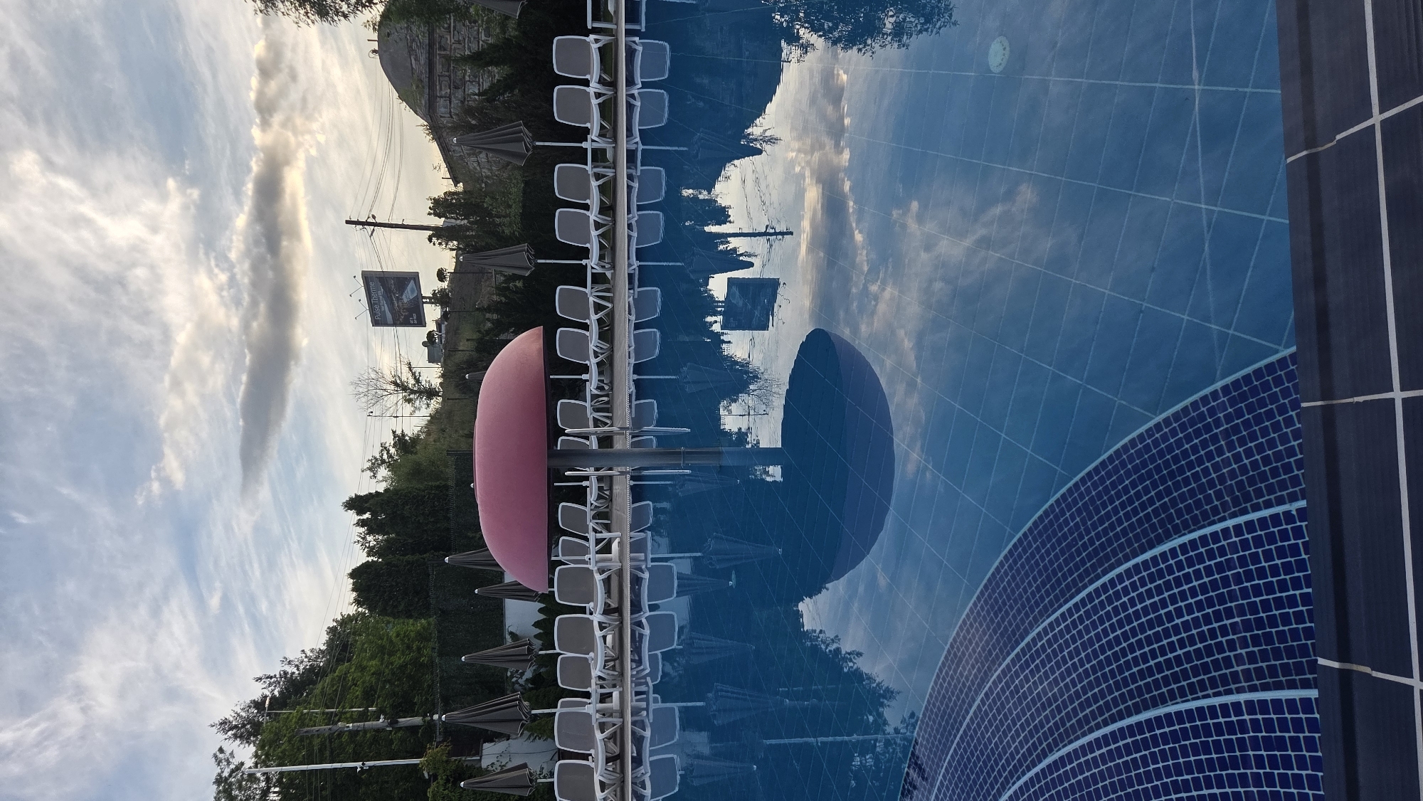 The image depicts an outdoor swimming pool area under a cloudy sky. In the foreground, the blue tiled edge of the pool curves inwards, with dark tiled coping bordering it. The water in the pool is calm and reflects the sky, surrounding trees, and a prominent red mushroom-shaped umbrella in the center. Behind the pool, a row of empty lounge chairs and closed umbrellas are neatly arranged on a paved area, backed by a dense line of trees and shrubs. In the background, a hillside covered in trees rises, with a road and utility poles visible. Further in the distance, a billboard with the text "Pulse Therme" is situated on a hill, alongside some buildings and more trees. The lighting suggests it is either early morning or late afternoon. No people are visible in the scene.