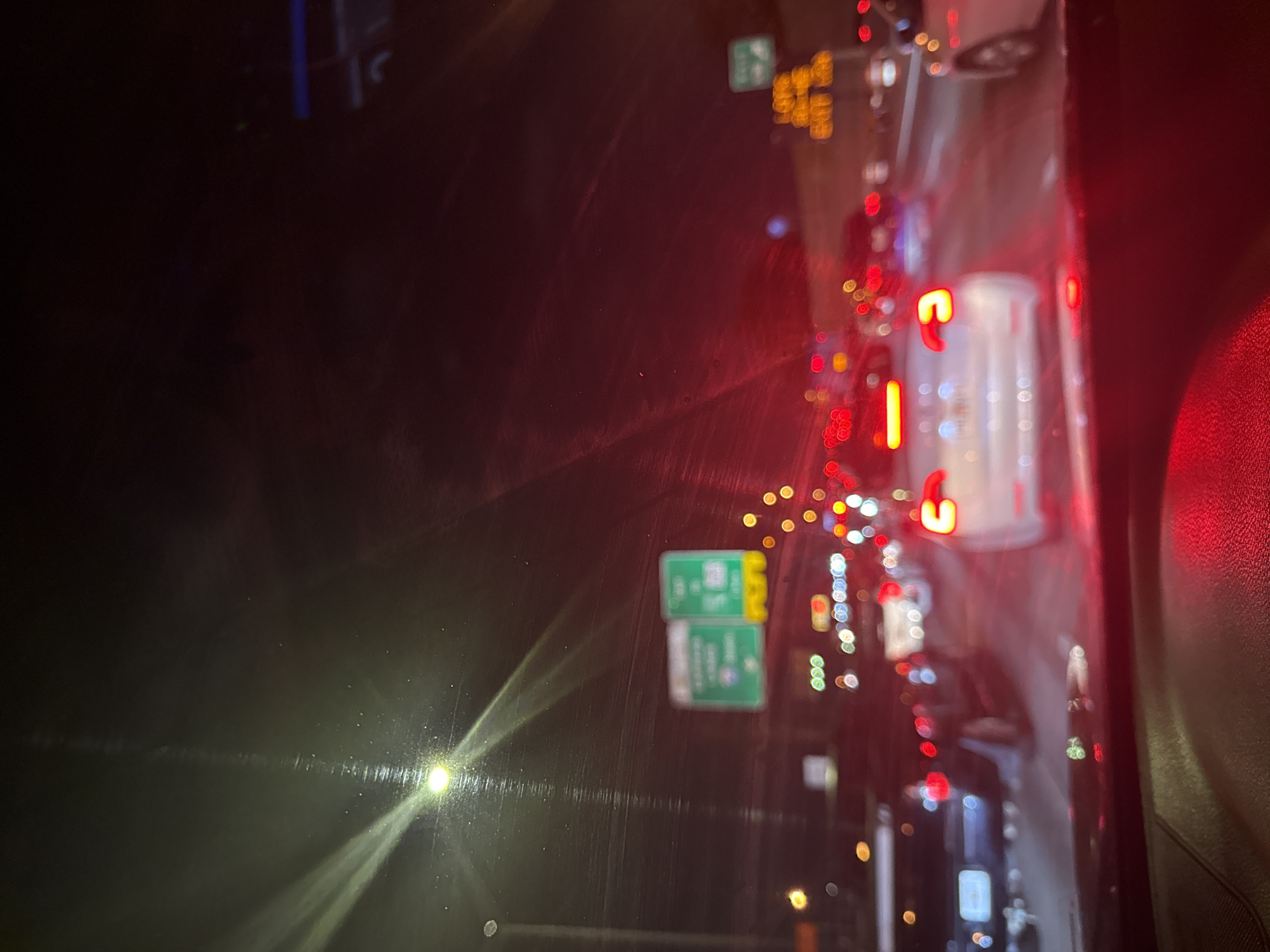 The image shows a view from inside a car, likely on a highway in North Miami, United States, at night. Traffic appears congested, evidenced by numerous red taillights and blurred headlights. Road signs are visible, including green exit signs, suggesting the presence of highway exits. The view is partially obscured by the car's windshield, which shows some light reflections and possibly raindrops. The scene depicts a typical nighttime traffic scenario.