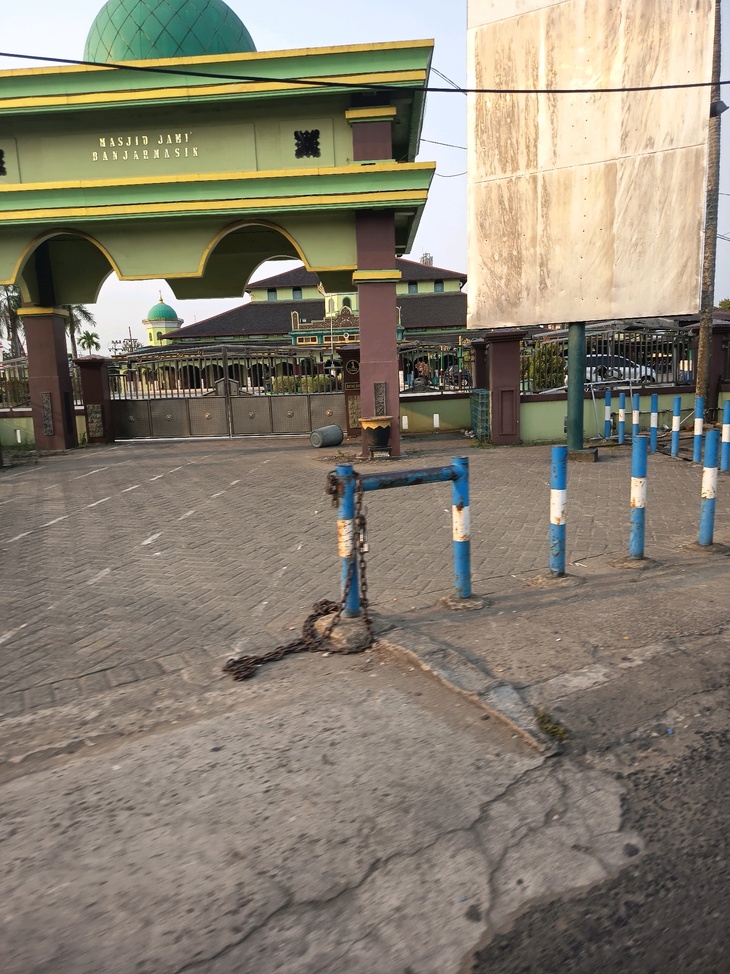 The image shows the entrance to Masjid Jami Banjaramsin, a mosque located in the city of Banjarmasin, Indonesia. The mosque features a prominent green dome and an arched gateway with gold lettering that reads "MASJID JAMI BANJARAMSIN". The gateway is flanked by pillars and decorative elements. In the foreground, a chain secured to a blue metal post is visible across a paved pathway, suggesting a temporary barrier or closure. Several blue and white striped bollards are also present, lining the entrance. A large, weathered white billboard stands to the right of the gateway. The background reveals more of the mosque's architecture and a metal fence. The scene appears to be outdoors, with clear sky and sunlight indicating it is daytime.