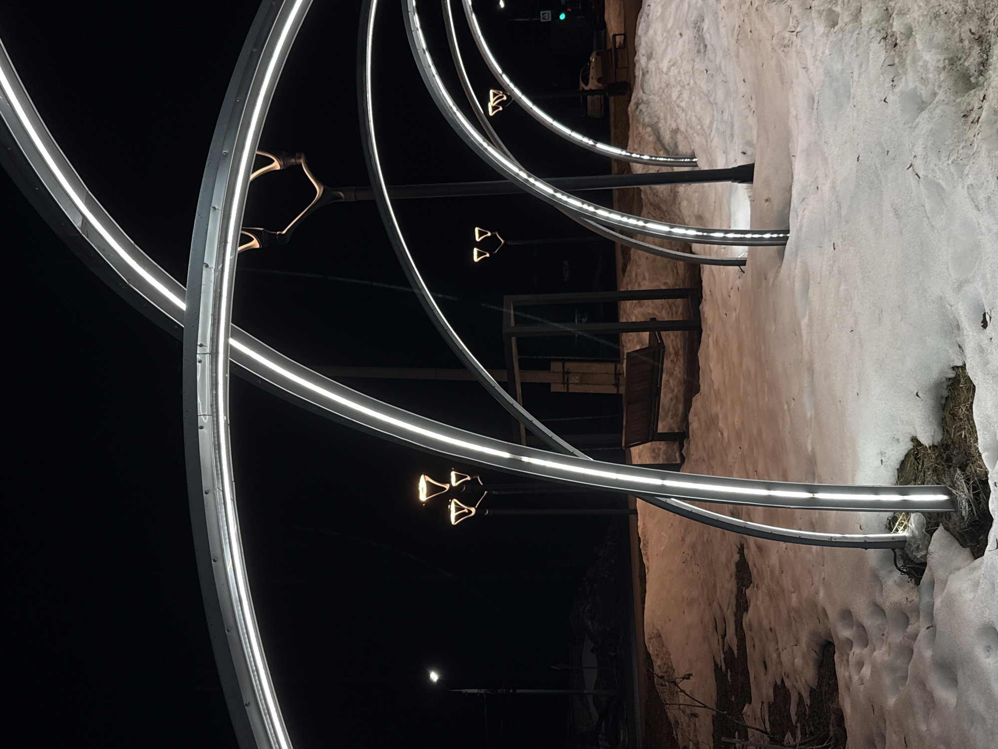 The image captures an outdoor scene at night in Chaikovsky, Russia, centered on an illuminated pedestrian pathway. Five dark metallic archways, each fitted with a continuous white LED strip light along its inner curve, dominate the foreground and mid-ground, creating a tunnel-like arrangement. These arches are anchored in ground covered with irregular patches of snow and exposed brown earth, interspersed with sparse dry vegetation.
In the mid-ground, two distinctive streetlights with tripod-like fixtures, each supporting three upward-glowing, inverted triangular light sources, illuminate the area with a warm hue. A standard, single-fixture streetlight is visible further in the background on the far left. Two dark wooden benches are positioned within the scene; one is visible to the left, partially obscured by an arch, and another is further back on the right. Beyond the arches and benches, a paved surface, likely a road or sidewalk, is visible. A white vehicle is partially discernible on the right side of this paved area, adjacent to a green traffic light. No individuals are present in the scene.
