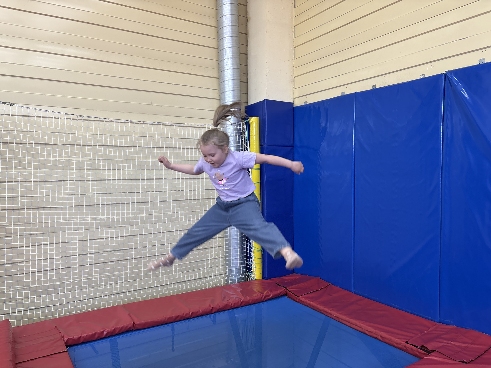 A young girl is jumping on a trampoline. She is wearing a purple t-shirt and grey pants. Her arms are outstretched, and her legs are spread apart. The trampoline has a blue surface with a red padded border. Behind the trampoline is a blue padded wall. To the left is a silver ventilation pipe. The walls are painted beige with horizontal grooves. A white net is visible behind the girl. The scene appears to be indoors, possibly in a recreational facility. The location is Unknown, Unknown.