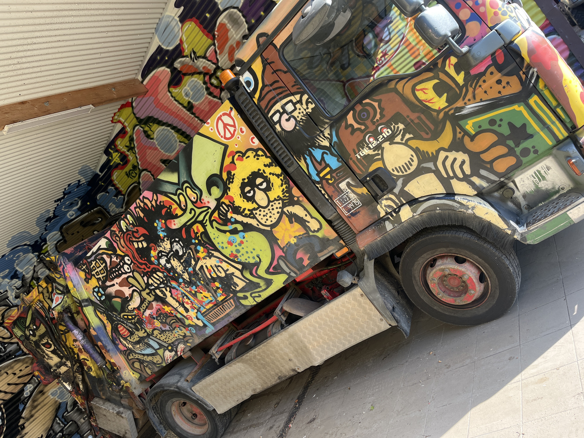 The image shows a vibrantly graffitied truck parked on a paved surface, likely a street or lot. The truck is adorned with a colorful and complex graffiti artwork featuring various cartoonish characters, abstract shapes, and bold lines. The graffiti covers the entire visible surface of the truck, from the cab to the cargo area. In the background, a corrugated metal structure is visible, possibly a building or a wall, also partially covered in graffiti. The lighting suggests it is daytime, with shadows cast on the ground. The truck appears to be stationary, with no people actively interacting with it. The overall scene is one of urban art and visual expression. While the image is provided without specific location context beyond the request for Copenhagen, Denmark, the style of graffiti and urban setting are characteristic of such a city.