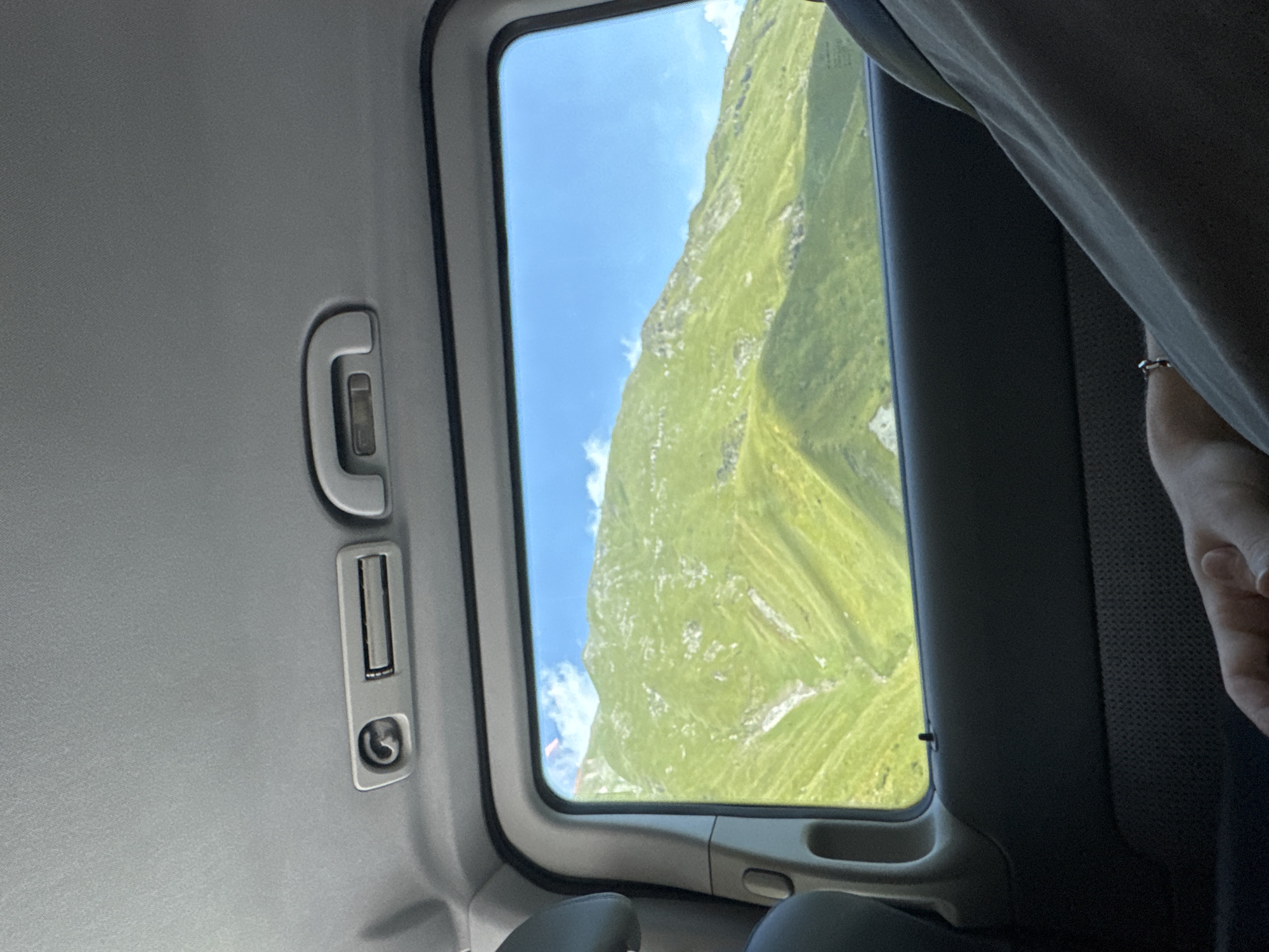 The image is taken from inside a vehicle, looking out of a window at a mountainous landscape. The mountains are covered in lush green grass, with some rocky outcrops and visible ravines. The sky is a clear blue with scattered white clouds. Inside the vehicle, the grey headliner of the car is visible at the top of the frame, with a light fixture and a handle. The interior of the car door is dark grey. A person's arm and hand are visible in the foreground, resting on a grey fabric surface, presumably a seat or clothing. The person is wearing a ring on their finger. The scene suggests a scenic drive through the mountains, possibly in or near Almasiani, Georgia, given the context provided, which is a mountainous region known for its natural beauty.