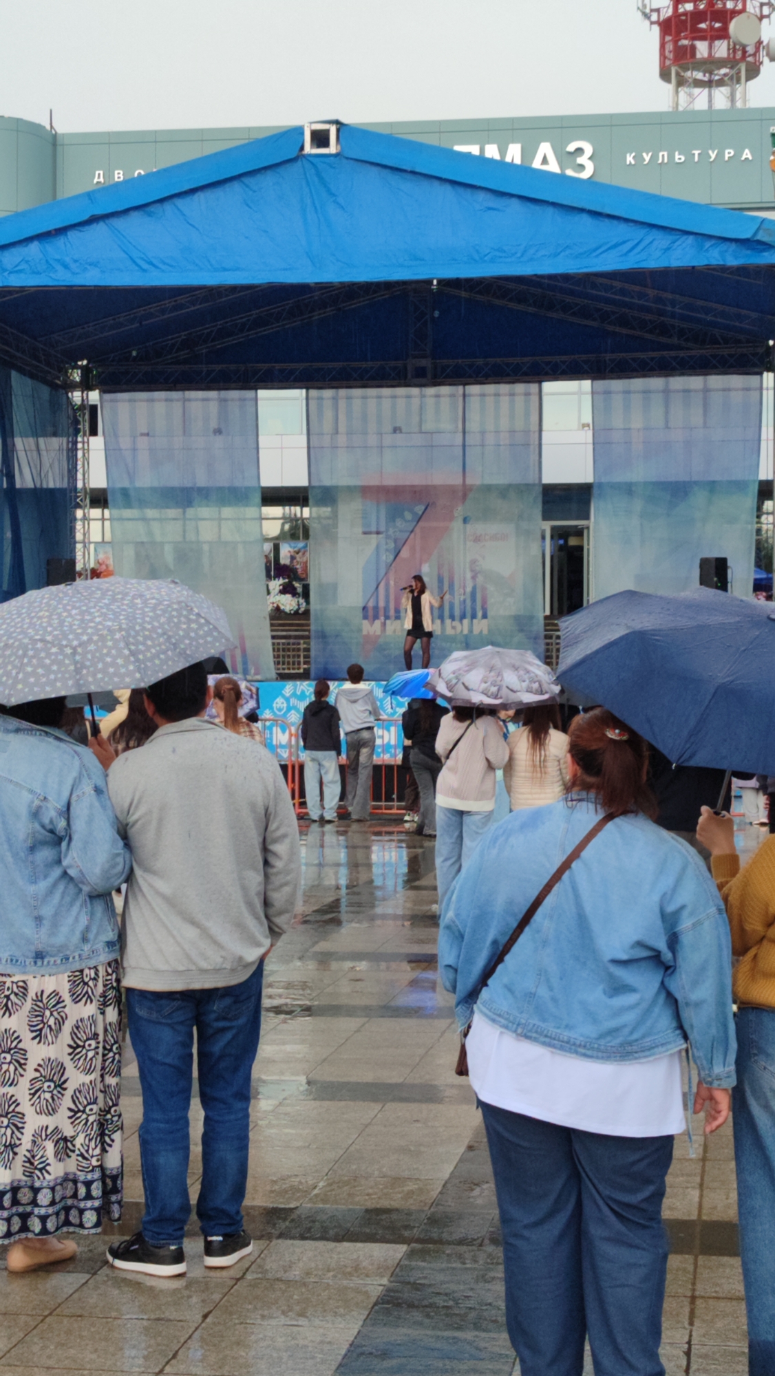A medium shot captures an outdoor event under a cloudy, rainy sky in the city of Mirny, Russia, likely at the Palace of Culture ALROSA, indicated by the building's signage. A blue tarp covers a stage where a female performer in a light-colored jacket and dark skirt sings into a microphone. A diverse crowd, many holding umbrellas, stands facing the stage, their reflections visible on the wet pavement. The foreground focuses on the backs of several attendees, showcasing casual attire like denim jackets, jeans, and patterned skirts. The scene evokes a sense of community gathering despite the inclement weather.