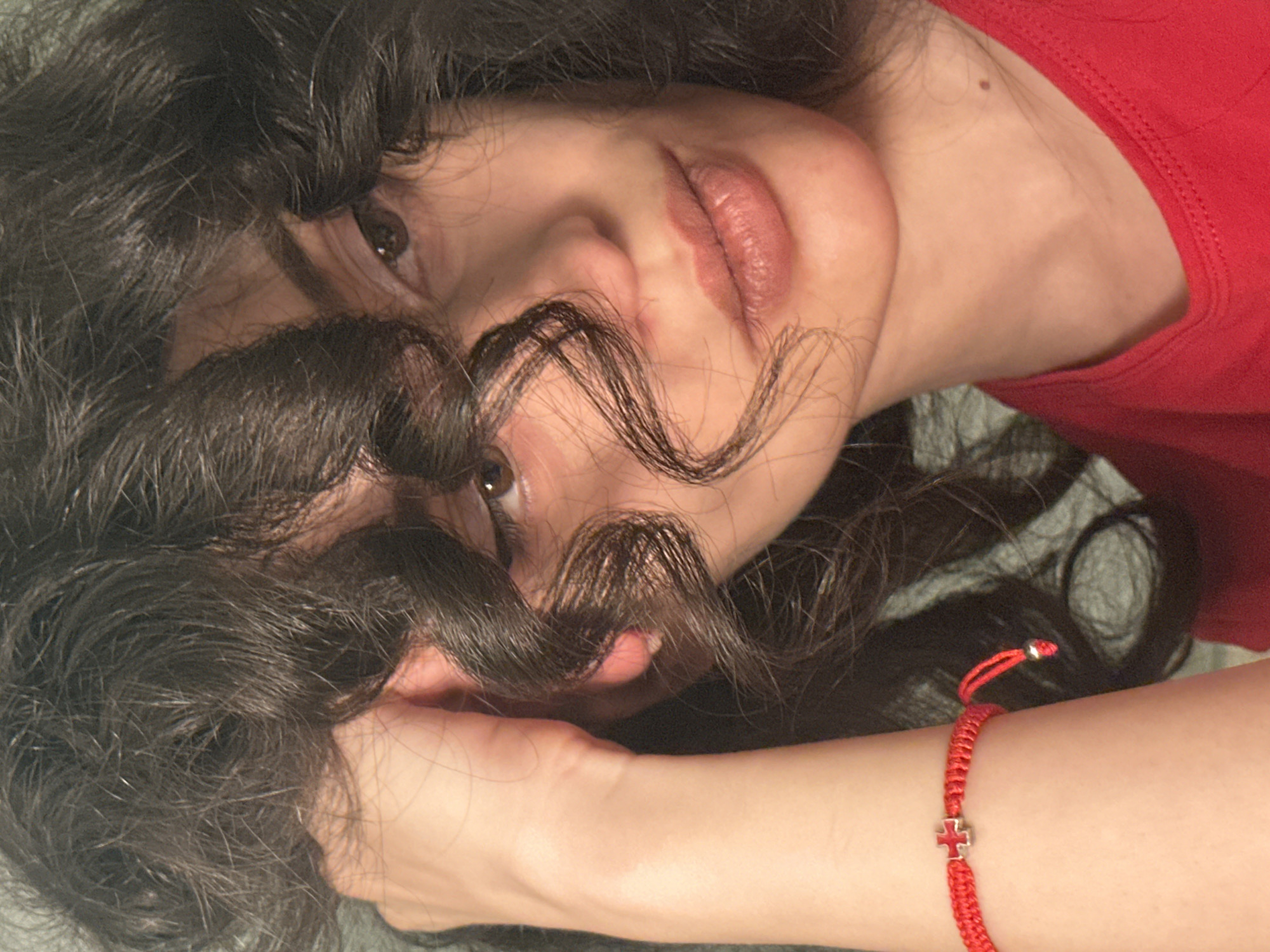 A close-up shot features a young woman with dark, curly hair. Her left arm is resting on her head, and her hand is partially obscuring her face. She is wearing a red shirt and a red string bracelet with a small, silver cross charm. The lighting suggests an indoor setting, possibly a home, with no discernible external city elements from Sofia, Bulgaria.