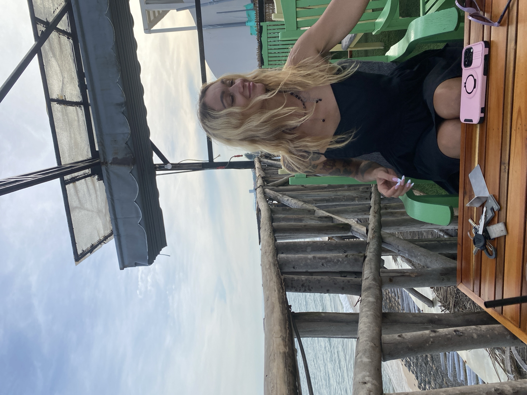 An overhead shot captures a blonde woman with wavy hair, wearing a black spaghetti strap dress, sitting in a green chair at a wooden table. Her eyes are closed, and she is smiling. A pink phone case rests on the table beside her. The background reveals a wooden fence overlooking the ocean and a cloudy sky. The scene appears to be a seaside cafe or restaurant in Phu Thuy Ward, Vietnam, likely during sunset due to the warm lighting.