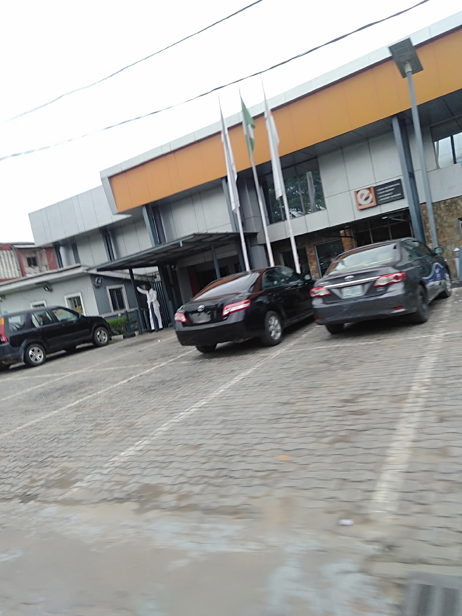 The image shows a parking lot with several cars in front of a building. The building has a light gray facade with a horizontal orange stripe near the top. There are multiple flagpoles in front of the building, with one flag appearing to be the Nigerian flag (green-white-green). The parking lot is paved with interlocking concrete pavers and has painted white parking lines. A black SUV is parked on the left side of the frame, and two sedans (one black and one dark gray) are parked closer to the building entrance. A person in a white suit stands under the building's entrance canopy. The location appears to be in a city in Unknown, Unknown.
