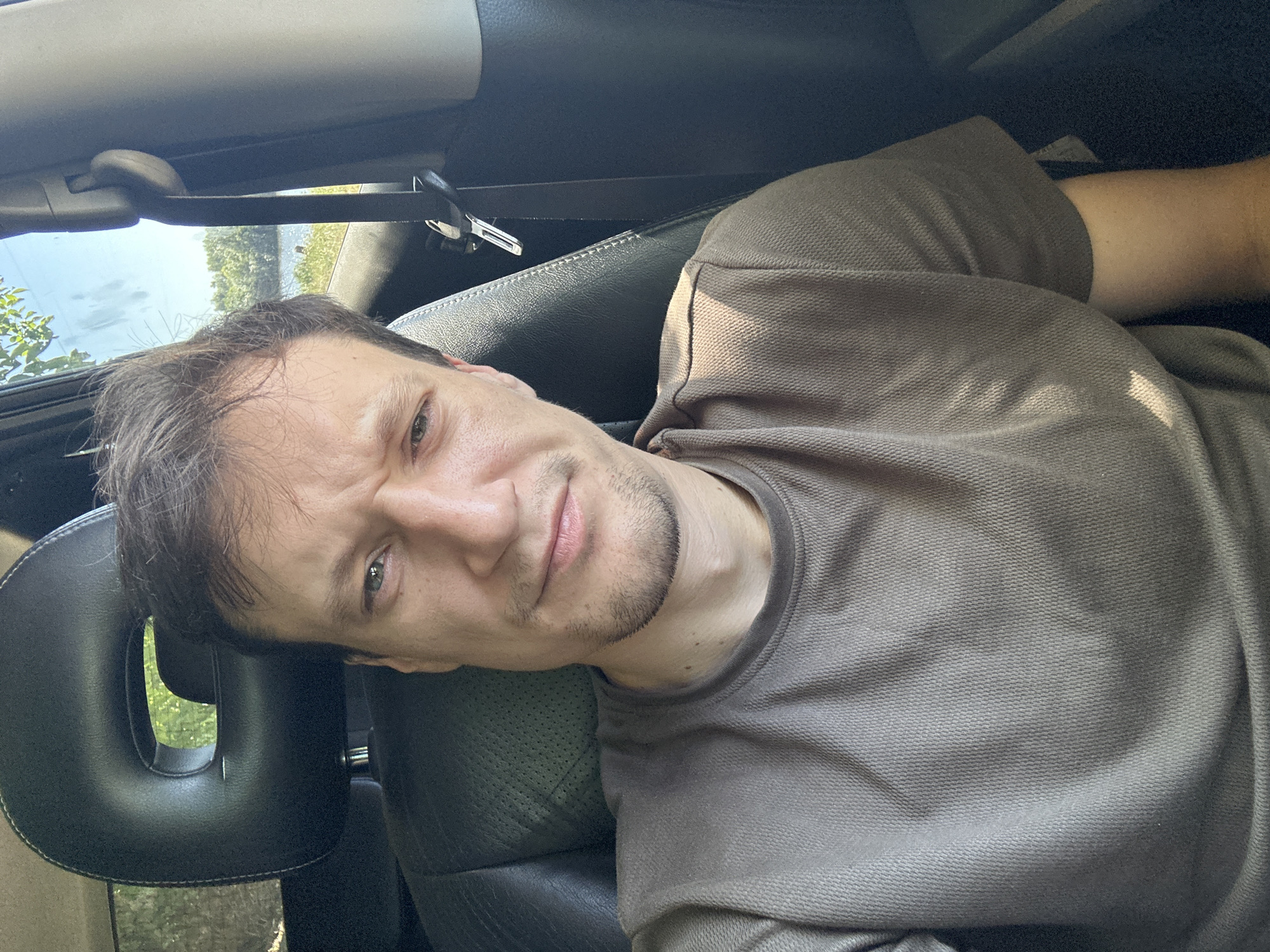 A man sits in the driver's seat of a vehicle, looking towards the camera with a slightly furrowed brow. The interior of the car is visible, with a black leather seat and headrest. Through the open window, a body of water and trees are visible in the background. The man is wearing a taupe t-shirt. The scene appears to be outdoors on a sunny day.
