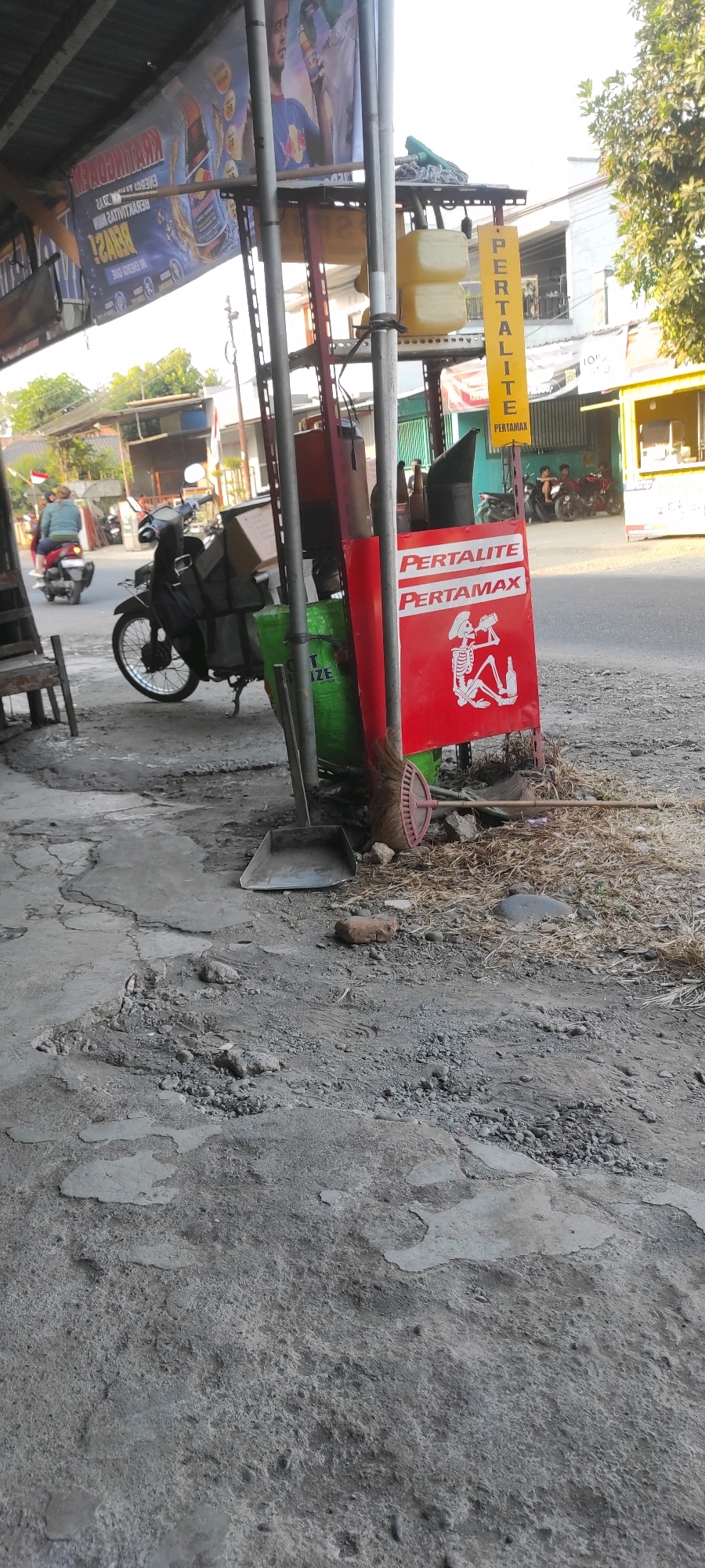 The image depicts an outdoor scene in what appears to be a roadside stall in Purwodadi, Indonesia. In the foreground, a rough, cracked concrete ground is visible, littered with debris, a metal dustpan, a brick, and dry straw-like material. Behind the ground, a motorcycle is parked, laden with items covered in dark grey fabric. A red sign is propped up, displaying the words "PERTALITE" and "PERTAMAX" with a graphic of a skeleton drinking from a bottle. To the left of the sign, a green cylindrical object is present. Further back, a yellow vertical sign with "PERTALITE" is visible. The background shows a street with vehicles, including other motorcycles and people, and buildings with a slightly faded aesthetic. Trees and foliage are also present. The overall impression is of a functional, everyday scene at a local establishment.