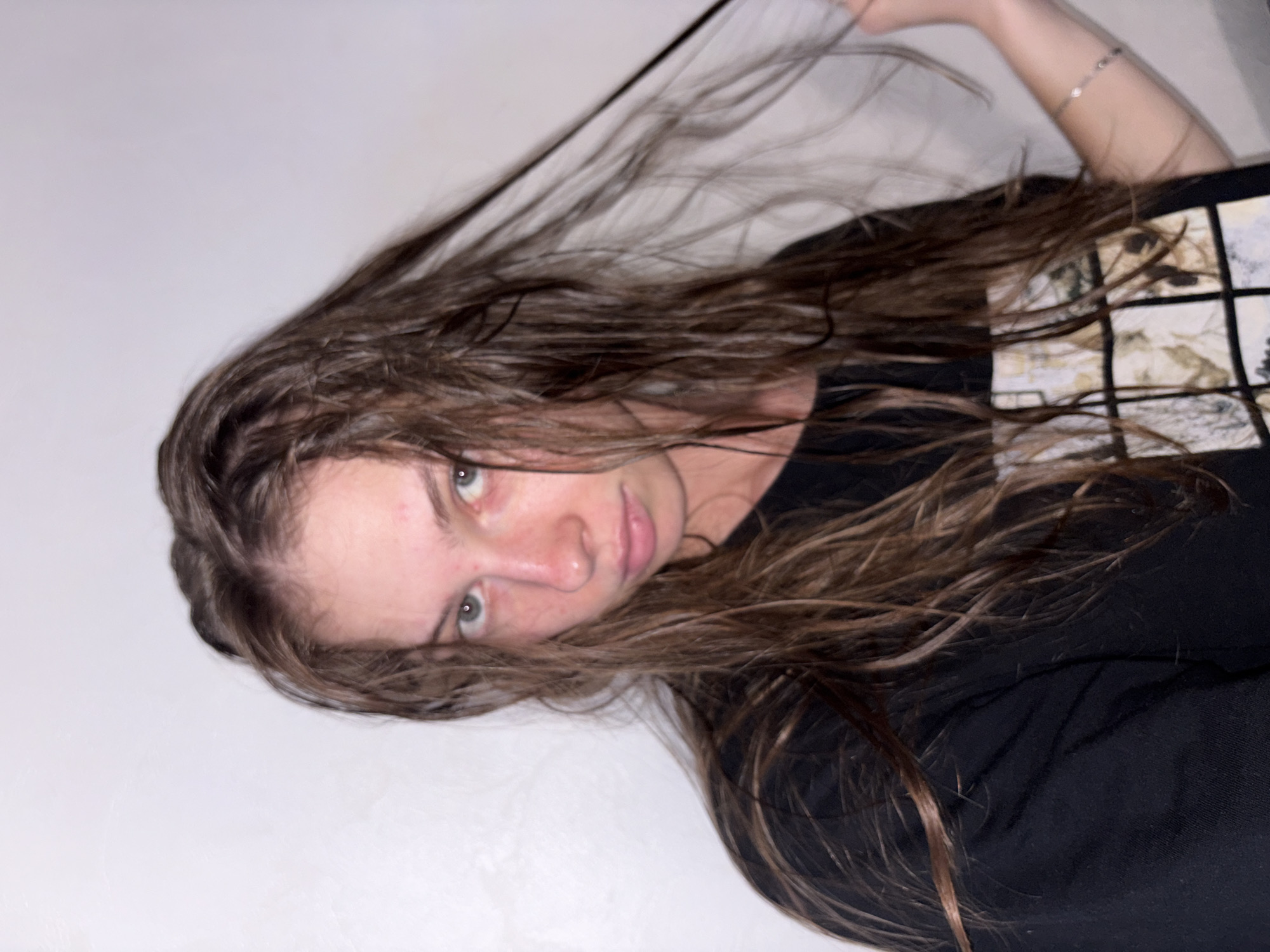 The media file shows a close-up, vertically oriented shot of a young person with long, wet, brown hair. Their face is tilted to the side, with visible eyes, nose, and lips. The person is wearing a black t-shirt with a graphic print on the upper right portion of the shirt. A thin, silver-colored bracelet is visible on their left wrist. The background is a plain, light-colored wall. The lighting appears to be artificial, casting some shadows and highlighting the wetness of the hair. The overall mood is casual and possibly relaxed, given the pose and wet hair, which might suggest a recent shower or activity involving water. There is no visible text in the image. The image appears to have been taken indoors.