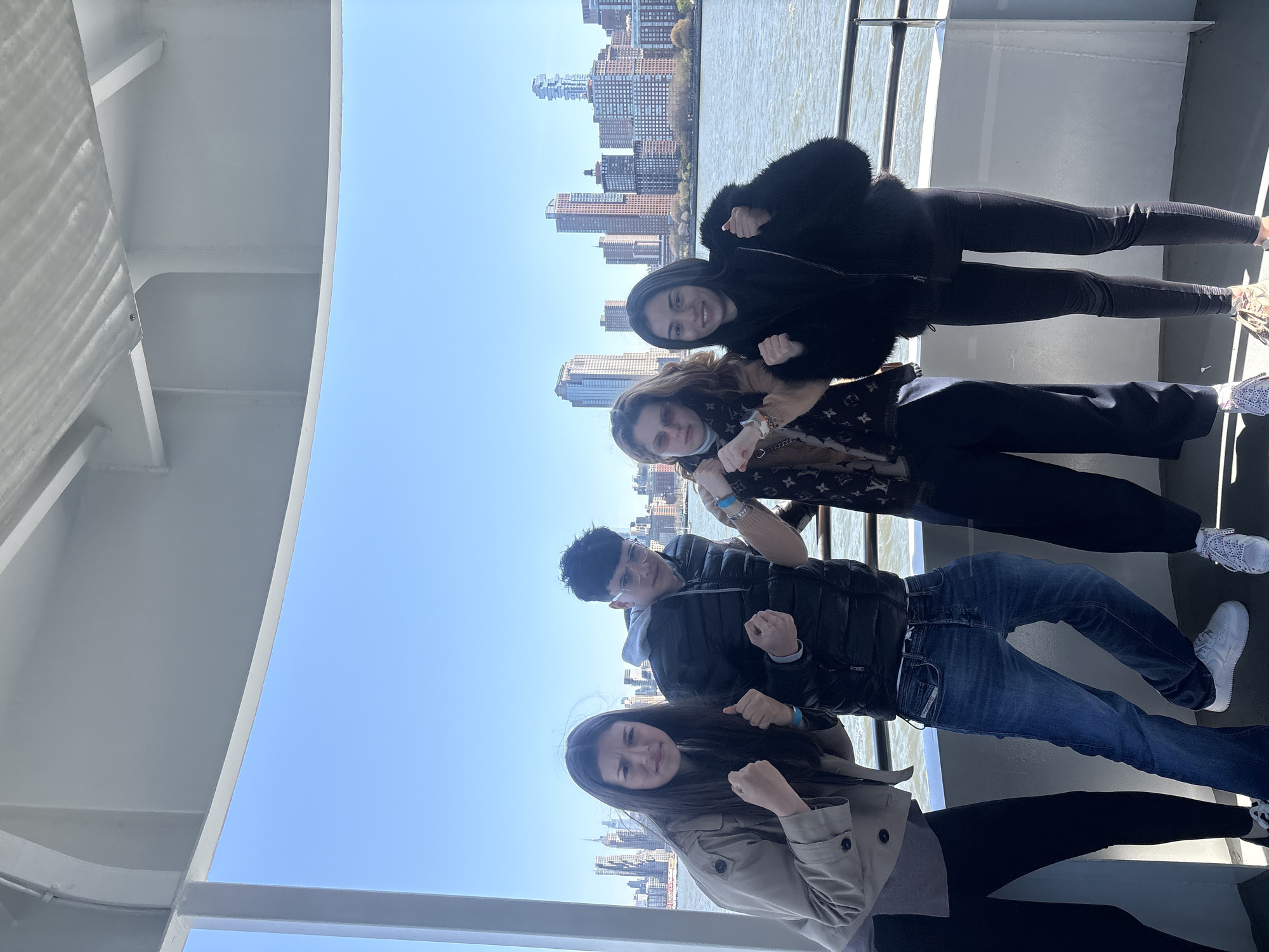 Four individuals pose for a photograph on what appears to be a boat or ferry. The backdrop features a cityscape with various buildings. The woman at the top is wearing a black fur-trimmed jacket and black pants. Below her, the second woman has long brown hair, sunglasses, a patterned Louis Vuitton-style scarf. The man in the middle wears a black jacket, glasses, and blue jeans. The woman at the bottom has long brown hair and is wearing a tan trench coat and black pants. All four individuals are clenching their fists and are posing to the camera. The shot is framed by what seems to be the metal frame of the boat. The location is Jersey City, United States of America, as evidenced by the buildings and the river.