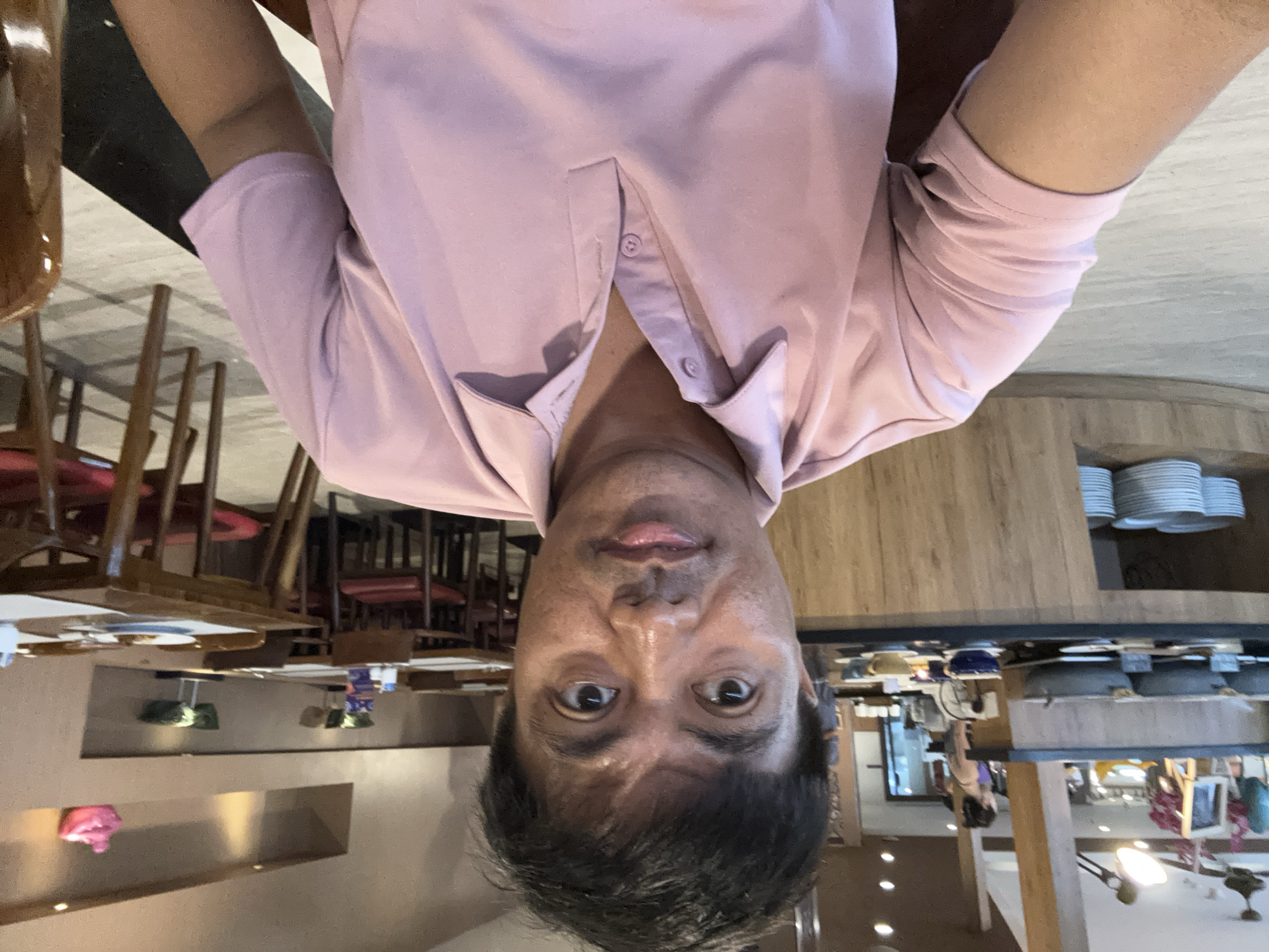 A selfie captures a man wearing a pink polo shirt in what appears to be a restaurant or buffet area. He is seated at a table and looking directly at the camera, with a blurred background that includes other tables and chairs, food service counters, and decorative elements. The setting suggests a public dining establishment, possibly within a hotel in Denpasar, Indonesia, given the provided context. The man's expression is neutral, and the lighting suggests an indoor environment with overhead illumination.