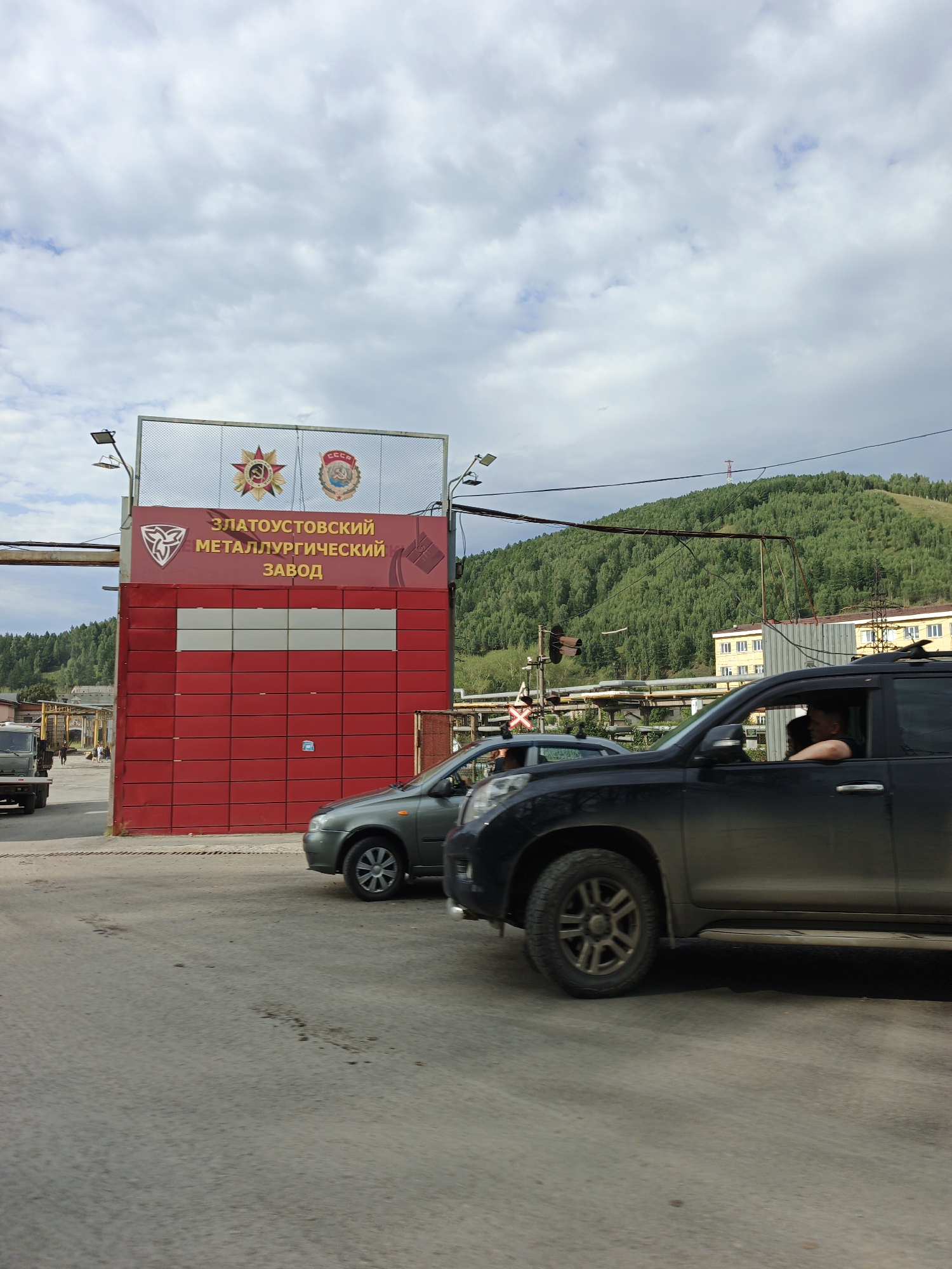 The image shows a bright red industrial building with a sign that reads "Zlatoust Metallurgical Plant" in Cyrillic letters. The building appears to be the entrance to a factory complex. Above the red building is a large metal sign with two Soviet-era insignia, one depicting a star with a hammer and sickle, and the other featuring a coat of arms with "CCCP" written on it.
In the foreground, two cars are visible on a paved road. A black SUV is in the right lane, with a person visible in the driver's seat. A smaller, grey sedan is in the left lane, also with a driver. The cars are stopped, possibly at a traffic light or railway crossing, as indicated by a signal pole with red and yellow lights and a red and white striped crossbuck sign visible to the left of the grey sedan.
In the background, a tree-covered hill rises behind the factory buildings. The sky is partly cloudy with a mix of white and grey clouds. The overall impression is an industrial area on a day with mixed weather conditions. The image was taken in Zlatoust, Russia.