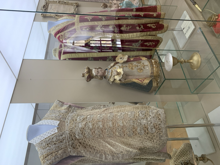The scene depicts an interior view of a museum exhibit at Nesvizh Castle, Nyasvizh, Belarus. Behind clear glass display panels, several historical and religious artifacts are presented.
On the left, a sleeveless, light gold-beige ceremonial garment or vestment is displayed on a headless mannequin. It features intricate silver-toned embroidery with floral and geometric motifs across its surface and a distinctive white, open-necked collar with a punched pattern.
In the center, a polychrome wooden statue of a crowned infant or child figure stands on a clear glass pedestal. The figure, possibly a representation of the Infant Jesus, wears a gold crown, a gold and white robe adorned with gold leaf accents and stylized floral patterns, and a dark blue or green cape. It holds a gold, multi-petaled object in its right hand.
To the right, a long, flowing red velvet or richly woven liturgical vestment, possibly a cope, is displayed on another headless mannequin. This garment is embellished with wide gold trim and large, repeating gold fleur-de-lis patterns. Reflections on the glass case in front of this vestment show parts of the surrounding room, including what appears to be a window or internal structure.
On a lower glass shelf in the foreground, directly below the statue, a brass or gold-colored chalice-like object with a flat, white disc (likely a paten) atop it is displayed. To the far left, a light-colored, woven or crocheted bag is visible on a small stand, partially obscured. The background features light-colored walls and a diffused, illuminated ceiling.