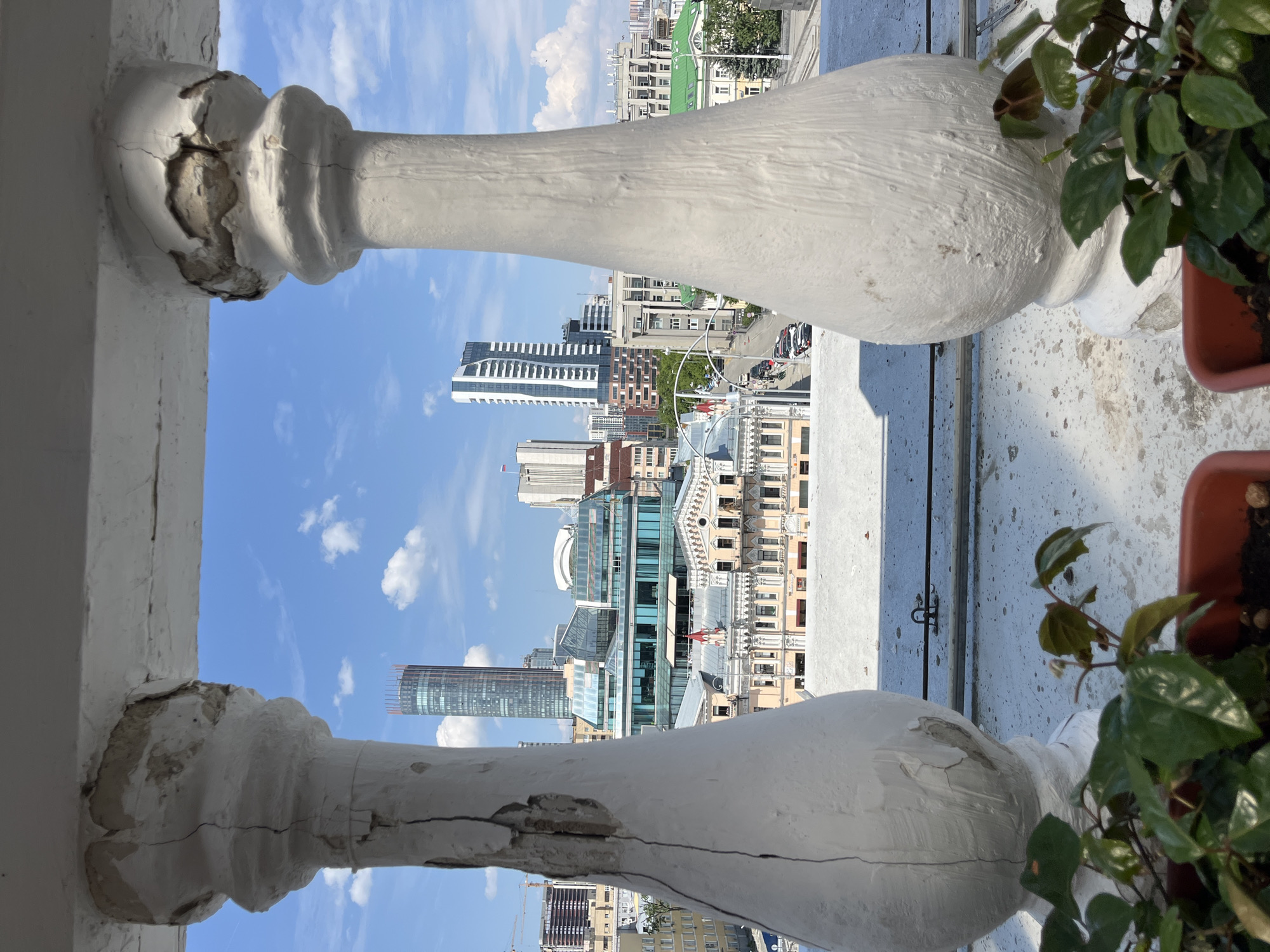 View of Yekaterinburg, Russia, from a balcony. Two heavily weathered white balusters frame the skyline. The balusters are cracked and chipped, revealing the inner material. Buildings of varying heights are visible, including skyscrapers. Clear blue sky with scattered clouds above. A potted plant with green foliage sits on the right side of the balcony.