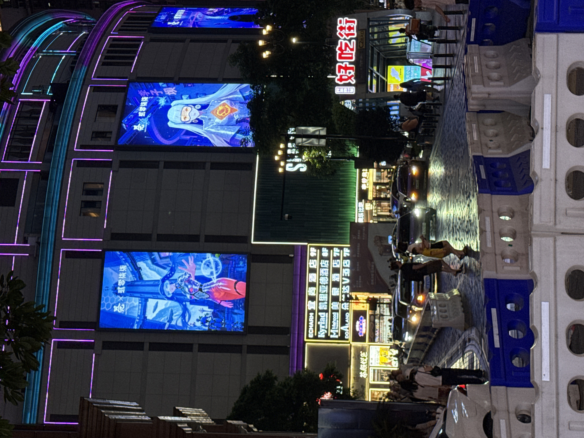 This image captures a bustling street scene at night in the Yuzhong District of China. The focal point is a large, modern building adorned with vibrant neon lights and massive digital billboards displaying animated characters. One billboard features a woman with long blue hair and a futuristic outfit, while another shows a different animated character in an orange and white costume. Below these, illuminated signs for hotels such as "ECHARM" and "Kyriad" are visible, indicating commercial activity.
The street level is characterized by flowing traffic, with headlights of cars illuminating the wet, cobblestone-like pavement. Pedestrians are seen walking along the sidewalk, some appearing to be tourists or shoppers. Temporary barriers, likely for construction or traffic management, are present in the foreground, dividing the street and sidewalk. The overall atmosphere is one of urban dynamism, with a blend of modern technology, commercial establishments, and everyday city life under the glow of artificial illumination.