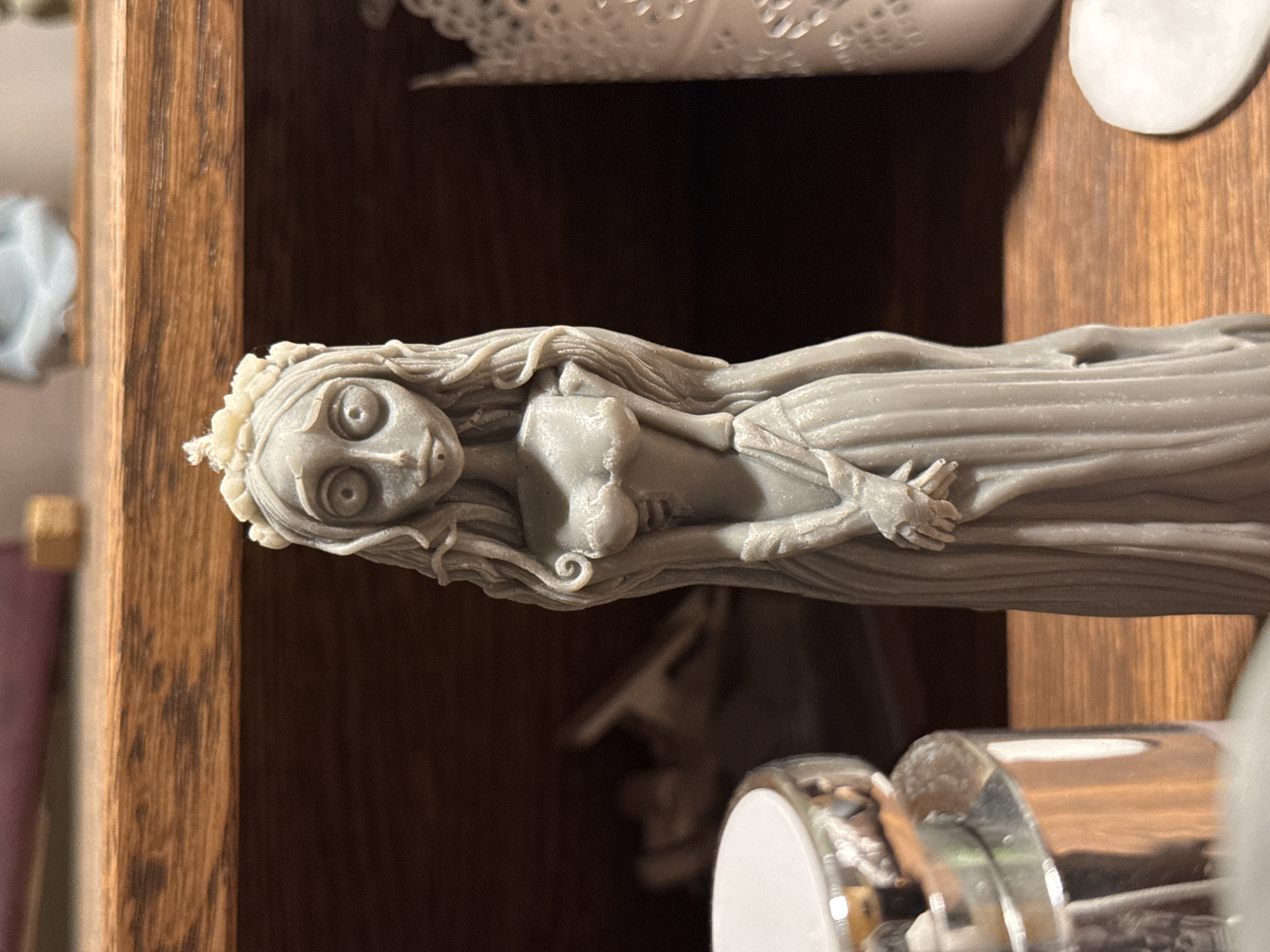 The image shows a close-up of a grey candle shaped like a figure with large eyes, long wavy hair, and a flowing gown. The candle is standing on a wooden surface, possibly a shelf. In the background, out of focus, are other objects, including what appears to be a white decorative item to the right and a metallic object with a white lid to the left. The overall setting appears to be indoors. There is no discernible information about the location in Бараўлянскі сельскі Савет, Belarus, within this image.
