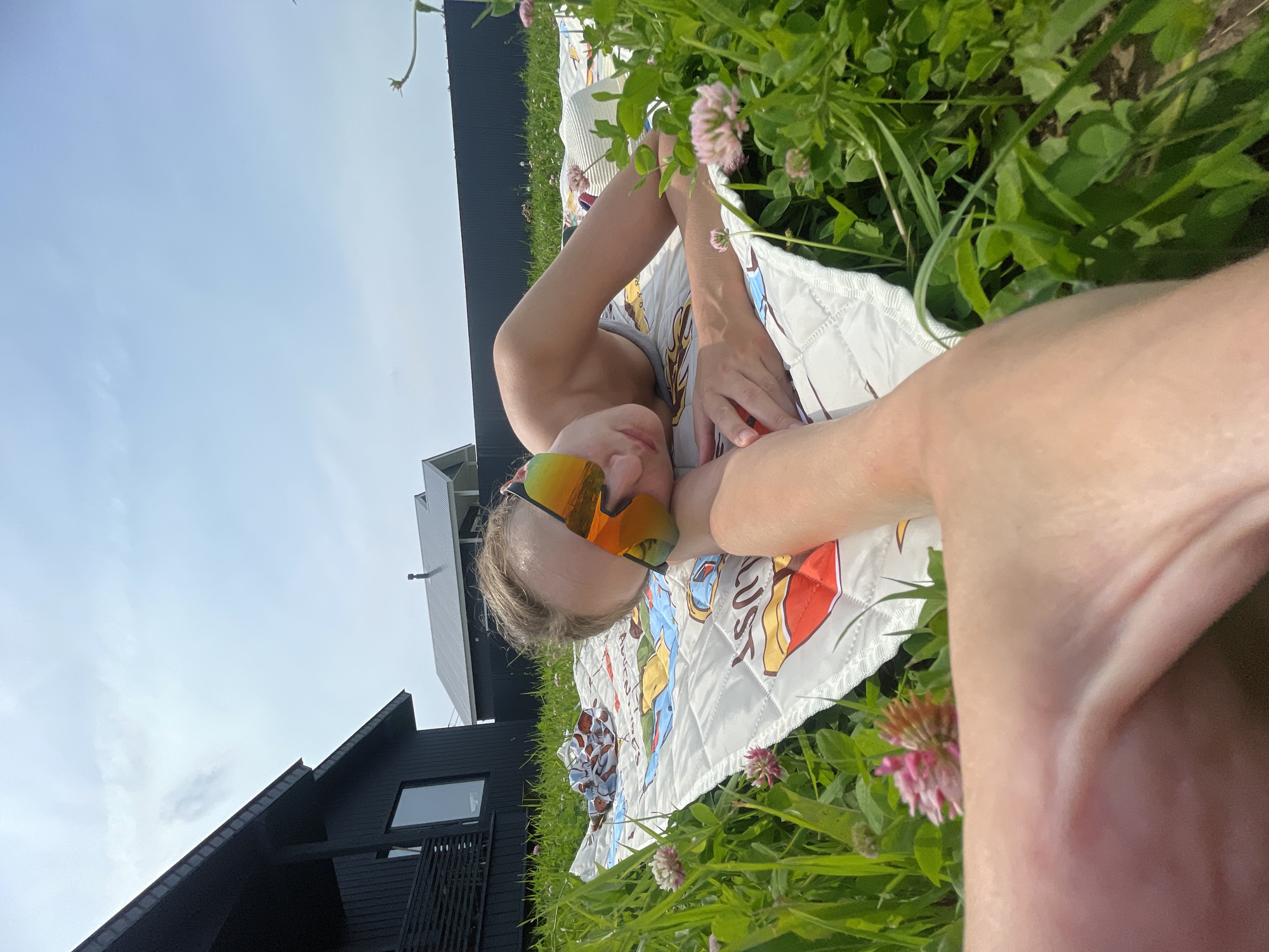 A person is lying on their stomach on a patterned blanket in a grassy area, wearing mirrored sunglasses. They are outdoors in what appears to be a rural or suburban setting, with a dark-colored house visible in the background to the left and another building and fence behind them. The sky is blue with some clouds. The grass is green and there are pink clover flowers growing. The foreground is dominated by a person's leg, out of focus. There is no indication this location is in Podshivalovo, Russia.