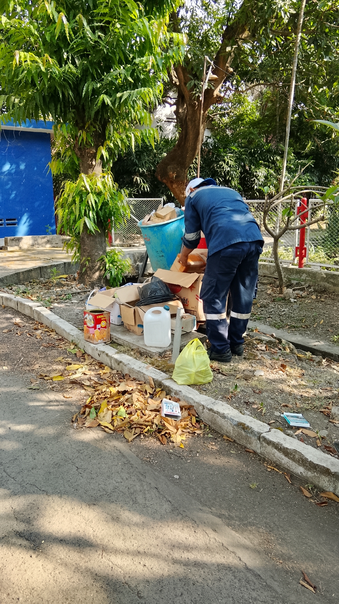 In Tlogobendung, Indonesia, a sanitation worker in a blue uniform and white cap is sorting through a pile of discarded cardboard boxes and other refuse next to a large blue trash receptacle. The scene is outdoors, with lush green trees providing shade. Fallen leaves are scattered on the ground, particularly near a concrete curb. Among the trash, there are several cardboard boxes, some open and empty, and one containing black plastic bags. A yellow plastic bag, a white plastic jug, and a red and gold cylindrical container are also visible. In the background, a blue building and a white fence suggest an industrial or institutional setting.