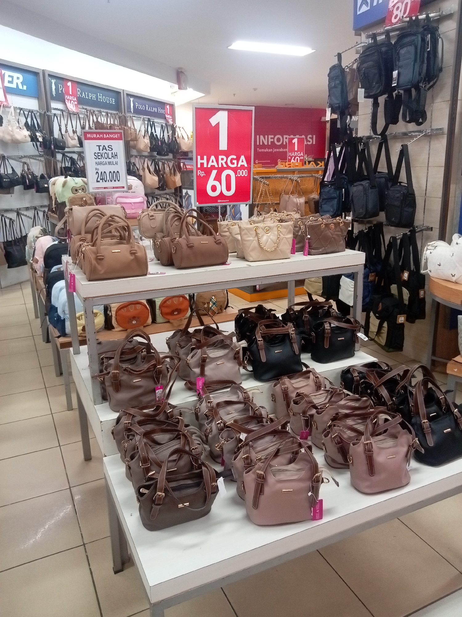 The image shows a retail display of handbags and backpacks in a store, likely located in Batam, Indonesia, indicated by the pricing in Indonesian Rupiah (Rp). The merchandise is organized on shelves and hanging racks. Prominent signage includes a red banner with white text indicating "1 HARGA 60.000" and another that reads "TAS SEKOLAH HARGA MULAI Rp. 240.000". A variety of bags are on display, including small to medium-sized handbags in neutral colors like beige, brown, and black, as well as some backpacks. The store environment appears to be well-lit, with tiled flooring and displays made of metal and wood. There are no people visible in the frame.