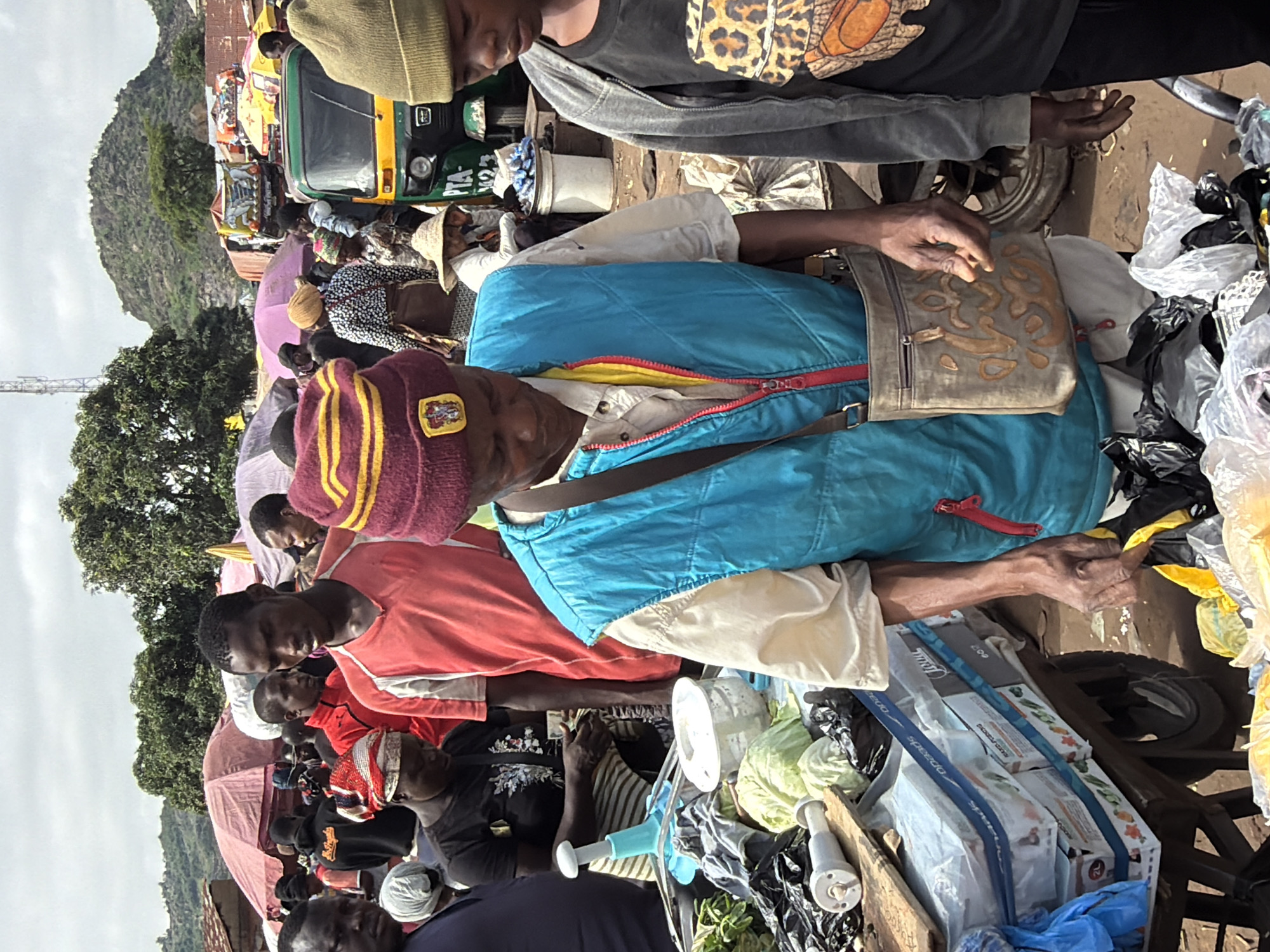 The image depicts a bustling outdoor market scene in Gura Top, Nigeria. In the foreground, a person wearing a blue vest and a maroon and yellow striped hat is examining items. To their right, another person in a grey hooded jacket with a leopard print design on their t-shirt stands. Behind them, a variety of market stalls are visible, with people browsing and interacting. Colorful umbrellas and tents provide shade. In the background, a green and yellow auto-rickshaw is parked, along with other vehicles and buildings. The ground is dusty and uneven. The overall atmosphere suggests a lively and active marketplace.