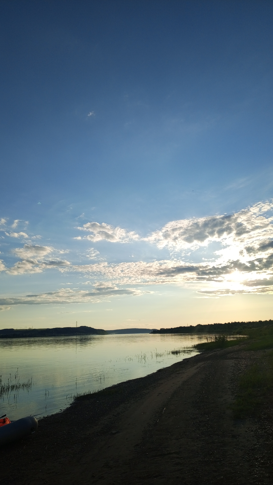 A wide-angle landscape photograph captures a tranquil riverine scene at either dawn or dusk in Шолья, Russia. The sky, occupying the upper two-thirds of the frame, transitions from a deep blue at the zenith to a lighter, paler blue near the horizon. Scattered cumulus clouds are present across the sky, becoming more dense and illuminated by a low-angle sun on the right side of the frame. A faint, light blue paraglider is visible high in the sky on the far upper left.
The midground features a broad, calm body of water, likely a river, reflecting the ambient light and sky in tones of pale green and blue. Sparse aquatic vegetation, primarily thin reeds, emerges from the water along the near shoreline. The foreground on the right depicts a dark, sandy or dirt path that traces the immediate edge of the water. On the far lower left, a portion of a grey and orange inflatable boat rests on the bank.
The distant horizon, across the water, reveals a landscape of low-lying hills or elevated terrain, covered with indistinct dark vegetation, likely trees. On the distant left bank, faint vertical structures, resembling power transmission towers or communication masts, are discernible against the sky. No human or animal subjects are explicitly visible, and no dynamic actions or interactions are occurring.