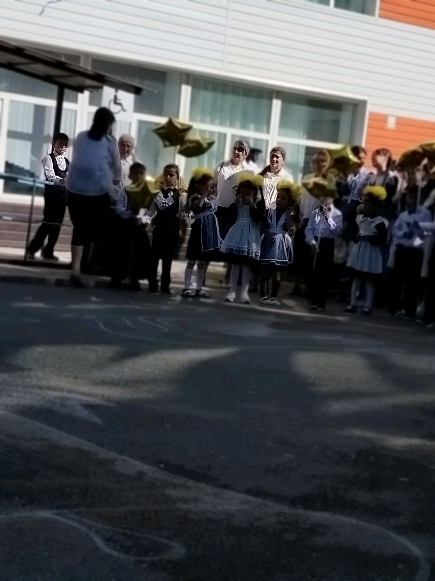 The image shows a group of people, predominantly children dressed in school uniforms, standing on an asphalt surface in front of a building. The children are holding star-shaped balloons. The building appears to be a school, with large windows and an orange-accented facade. A ramp with a wheelchair symbol is visible near the entrance. The overall scene suggests a school event or gathering, possibly a celebration or graduation, taking place outdoors in a city setting. The specific location is identified as Kölkent, Kazakhstan.