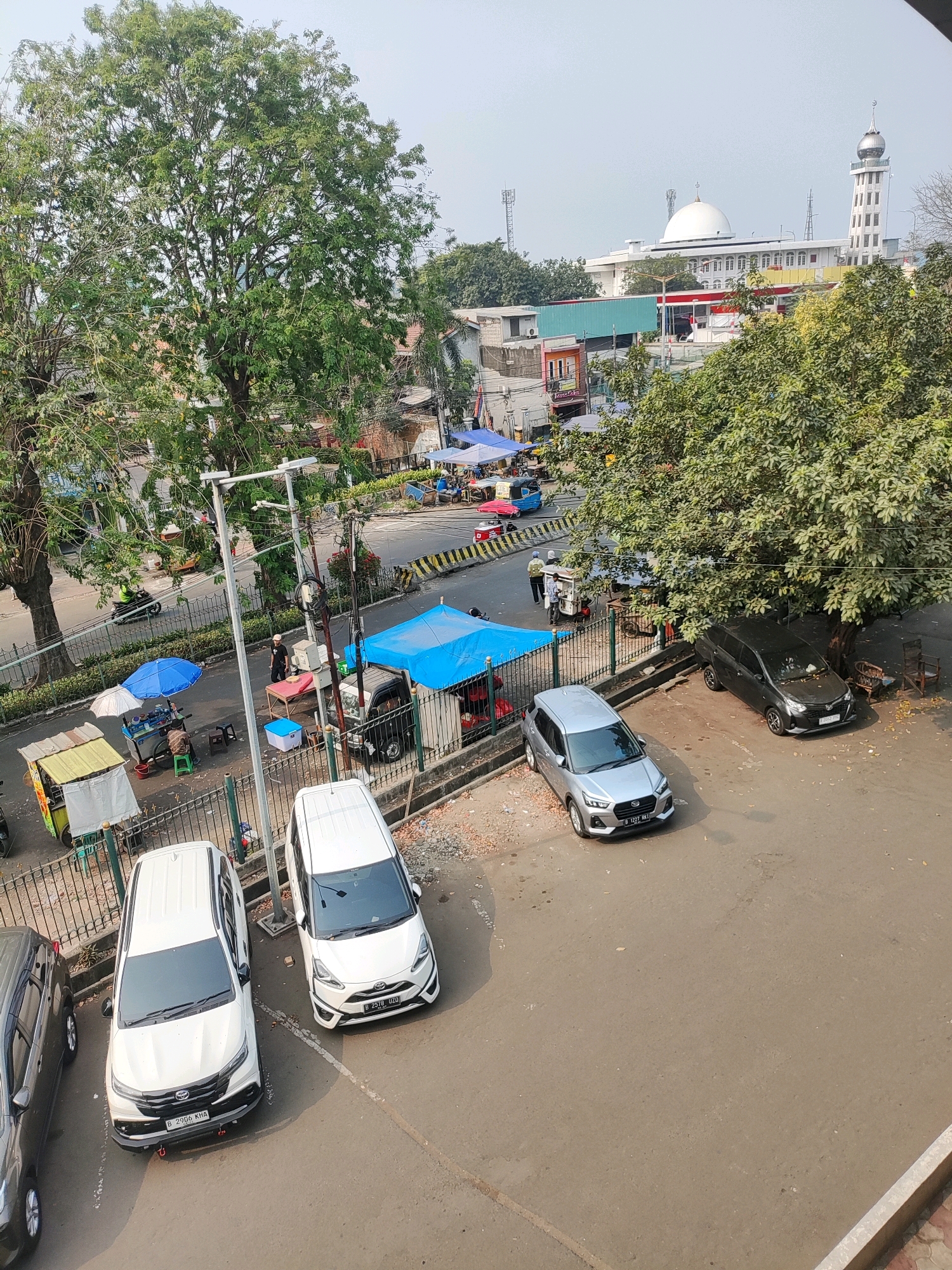 In East Jakarta, Indonesia, this image captures a street scene featuring a large tree, various vendors with blue tents and carts, parked cars, and a mosque in the background. The street appears busy, with at least one motorcycle visible and several people interacting with the vendors. The mosque, a prominent structure with a white dome and a tall minaret, is situated across the street from the vendors. The weather seems clear and sunny.