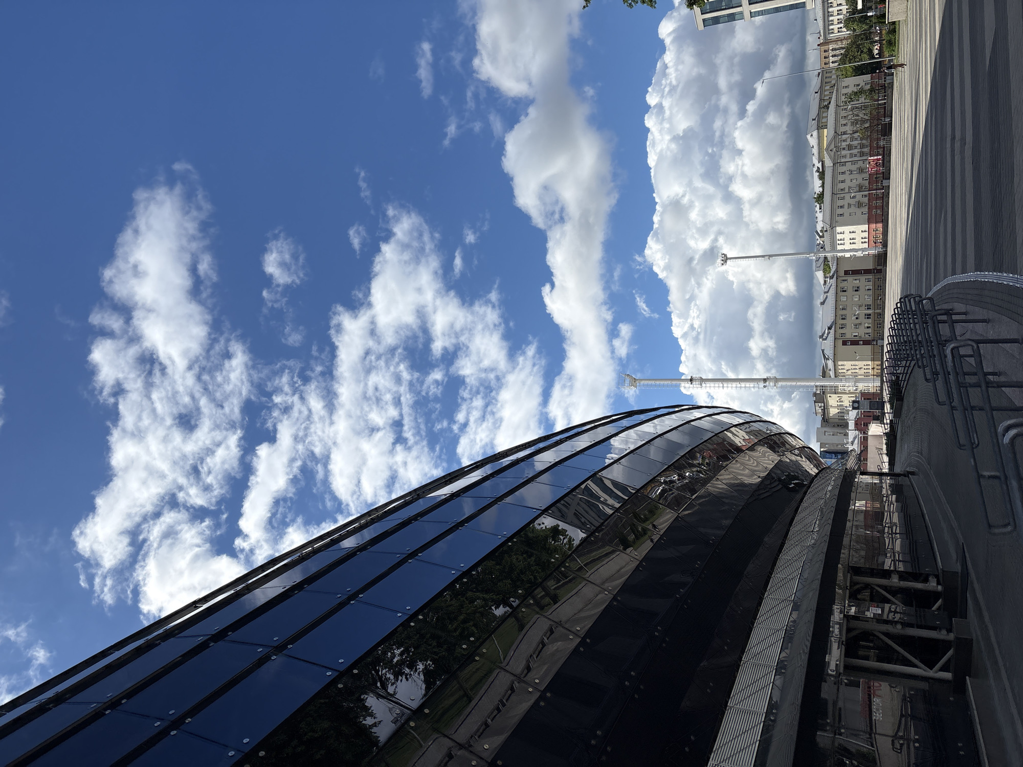 Here is a description of the image:
The image depicts a modern architectural structure located in Minsk, Belarus. The building features a curved facade constructed of dark glass panels. The glass reflects the surroundings, including trees, clouds, and other buildings, creating a mirrored effect. Above the building, the sky is bright blue with scattered white clouds. In the background, there are several buildings, a street, and some lampposts. To the right, there is a bike rack. The orientation of the photo is on its side.