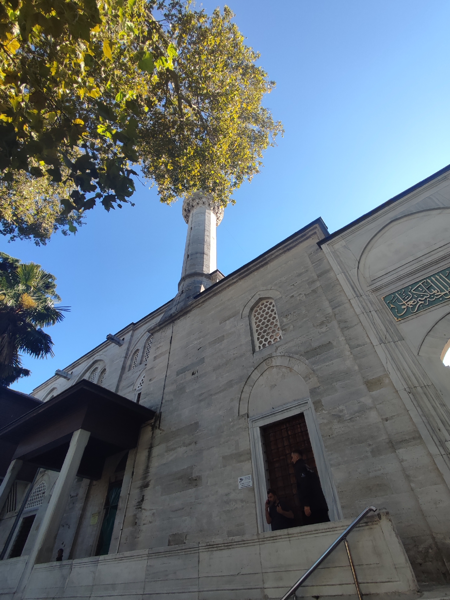 This is a low-angle shot of a mosque in Üsküdar, Turkey, featuring a minaret and a portion of the main building against a clear blue sky. A large tree with yellowing leaves shades the upper left of the frame, with its branches reaching towards the sky and obscuring part of the minaret. The mosque's facade is constructed of light-colored stone blocks, with arched windows featuring intricate latticework. A grand entrance with a wooden door is visible, and two men are standing in the doorway, one of whom appears to be on the phone. A metal handrail leads up to the entrance. In the background, a palm tree is partially visible on the left. The overall atmosphere is one of historical architecture set against a natural backdrop on a sunny day.
