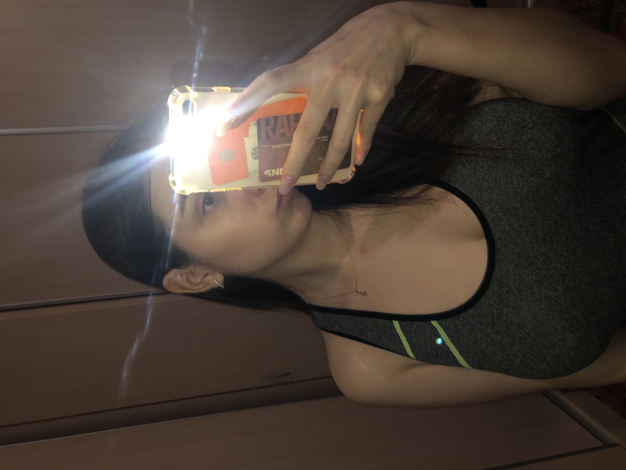 A young woman takes a selfie with a phone. The phone is held in her right hand, and the flash is on. She is wearing a gray sports bra with green stripes. She has a gold necklace on and a small hoop earring in her left ear. The background appears to be a wooden cabinet. There is no information suggesting the location is Noviy, Russia.