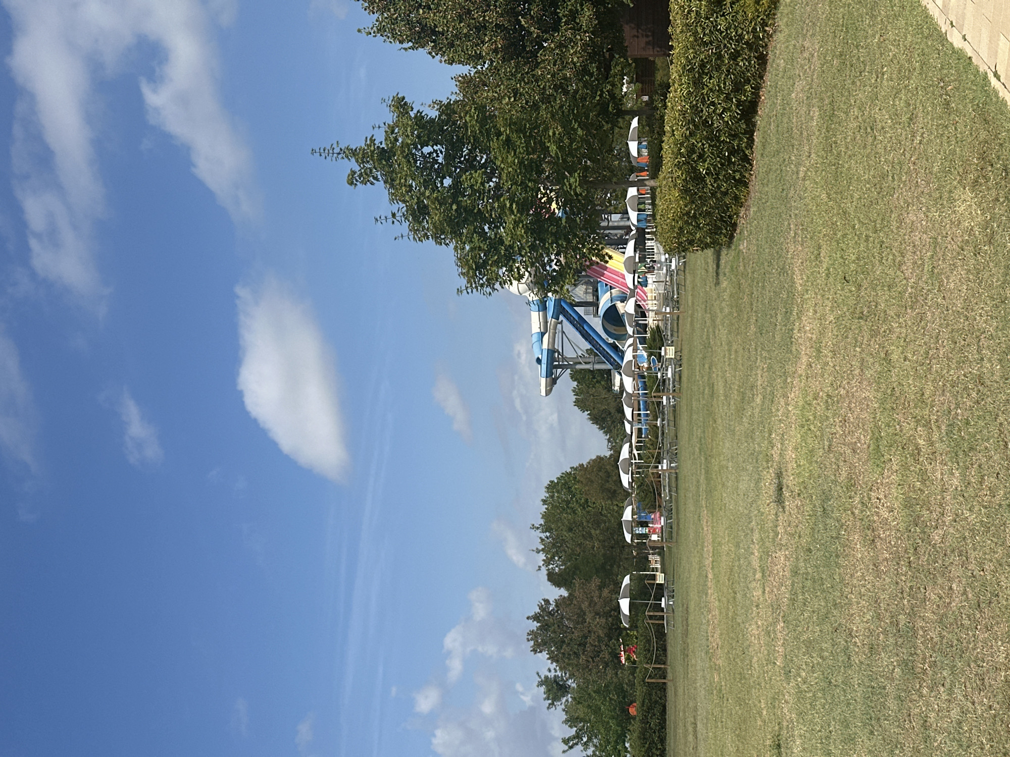 This is a photograph of a water park on a clear, sunny day. In the background, a large blue and white water slide with colorful striped sections is visible. In front of the slide, there are several rows of lounge chairs and umbrellas set up on a grassy area. Trees surround the park, and the sky is a bright blue with scattered white clouds. The foreground is dominated by a well-maintained lawn. The image does not provide information about specific people, actions, or interactions. The location in Ravenna, Italy, is not evident from the visual details alone.