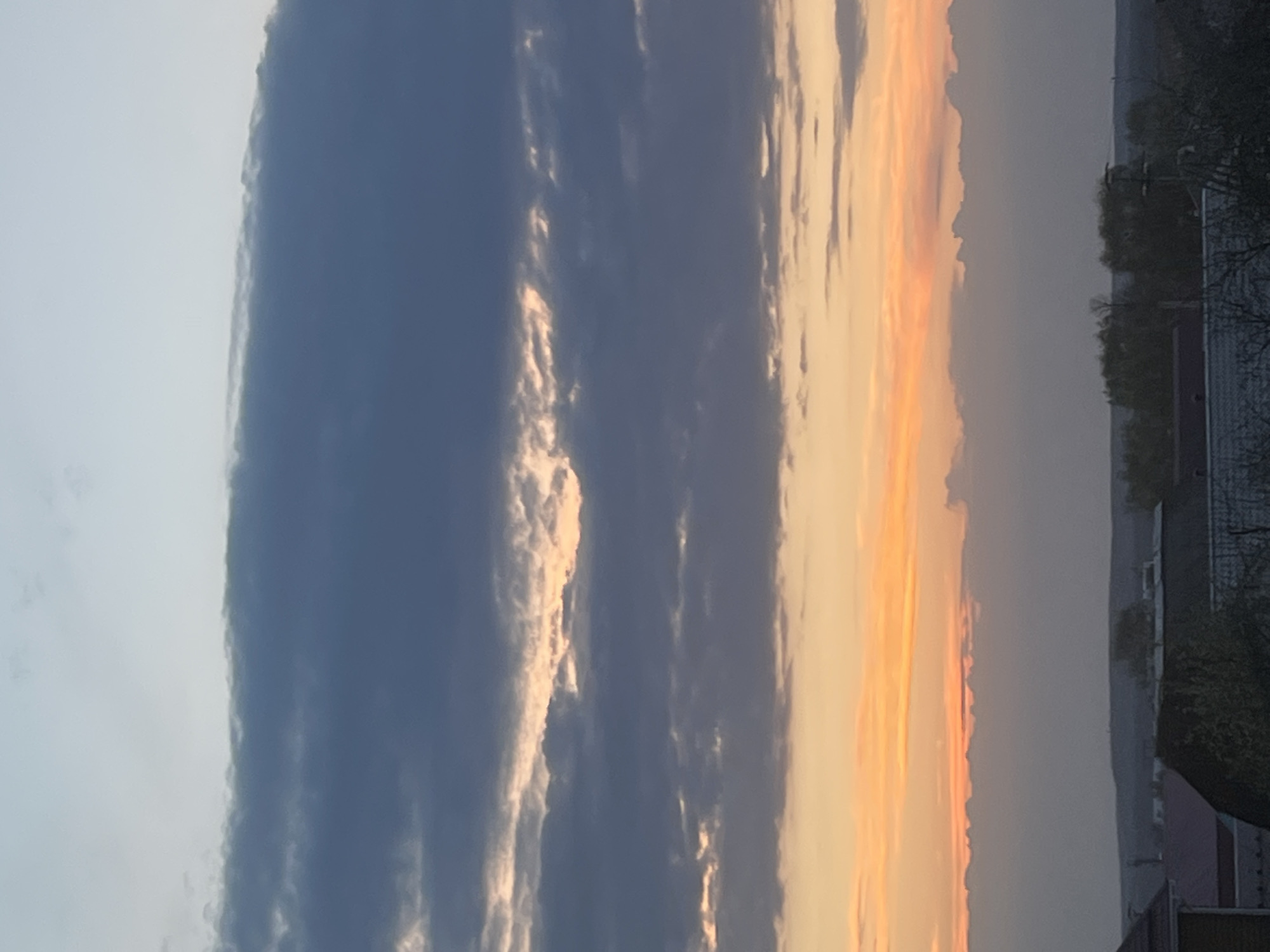 The image captures a sunset in Votkinsk, Russia. The sky is dominated by a combination of dark, cloudy bands and areas illuminated by the setting sun, creating an orange and yellow glow. In the foreground, rooftops, trees, and some indistinct building structures are visible, suggesting a residential or urban area. The overall scene depicts a tranquil moment as day transitions to night in the city.