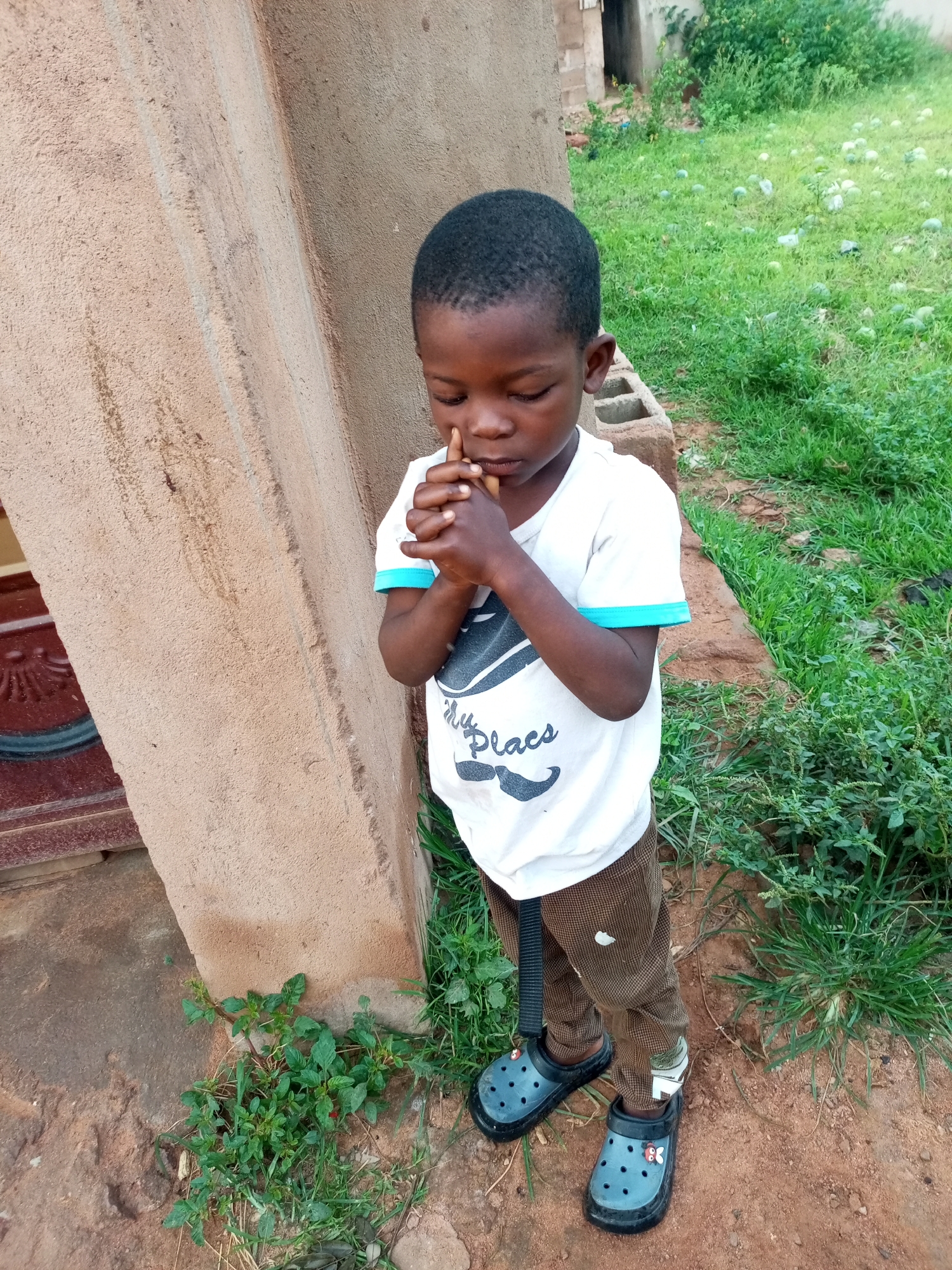A young Black child stands in front of a textured, light brown wall, facing slightly to the right and looking down with closed eyes and hands clasped in front of their chest. The child wears a white t-shirt with a graphic print on the chest and teal trim on the sleeves, brown corduroy pants, and dark blue Crocs-style shoes. The ground is a mix of red earth and green grass, with some scattered green foliage near the wall and in the background. In the background, there is more dense green vegetation and hints of some lighter green spherical objects scattered on the ground, potentially watermelons, and a rudimentary structure. The scene appears to be outdoors, possibly in a residential area, in the city of Gombe, Nigeria.