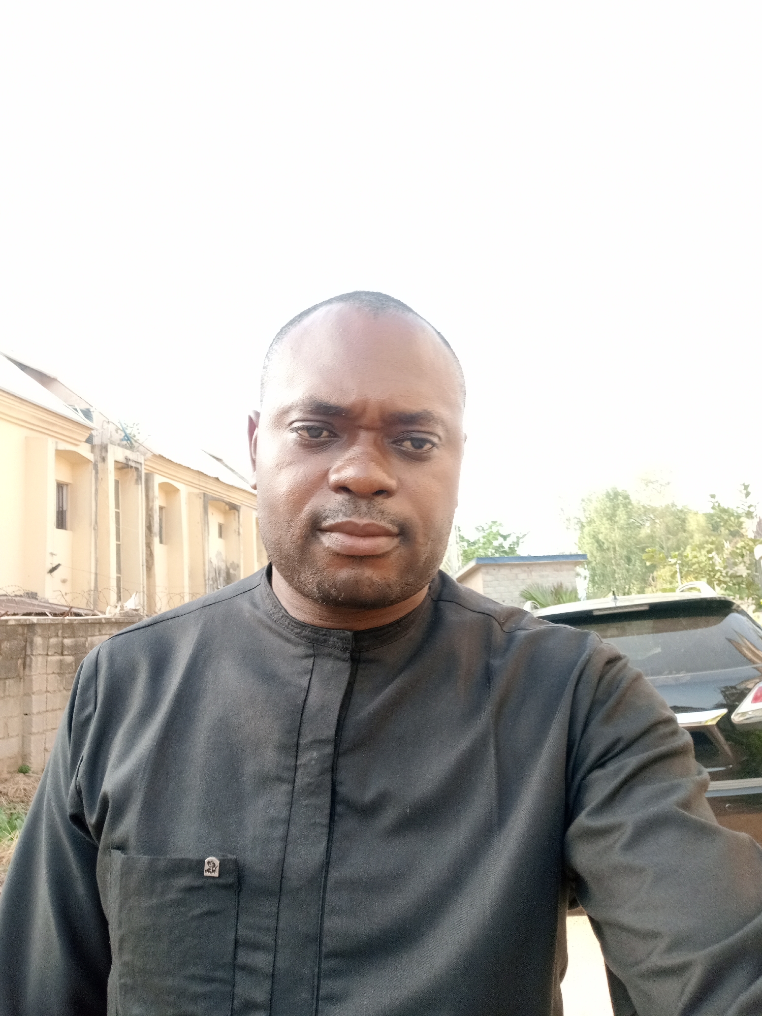 A man is captured in a close-up, eye-level shot, likely a selfie, with a bright outdoor background. He has dark skin, short dark hair, and a neutral, direct gaze towards the camera. He is wearing a dark (appears black or dark grey) long-sleeved, collared shirt, possibly a traditional African garment, with a chest pocket on the left side. A small, metallic, silver-colored emblem, possibly a stylized symbol or letter like an Omega, is attached to the flap of the pocket.
The setting appears to be an urban or semi-urban environment in Gwarinpa, Nigeria, during the daytime under a very bright, possibly overcast or overexposed sky, which appears almost white. To the man's left, a light-colored building with multiple windows and architectural details is visible behind a concrete wall topped with barbed wire. To his right, the rear portion of a dark-colored car (possibly an SUV or sedan) is visible, with green trees and some distant structures further in the background. The bright lighting casts a slight glare on the man's face and shirt.