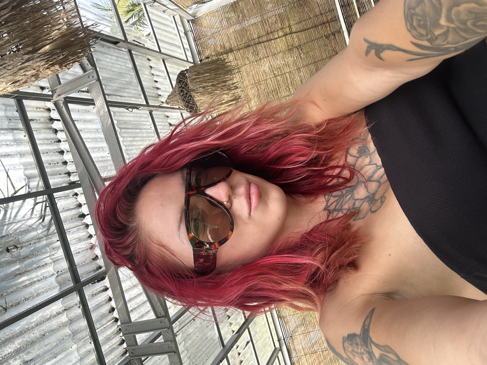 A selfie of a woman with red hair and tattoos wearing sunglasses, taken at the Nusa Penida Treehouse in Nusa Penida, Indonesia. The location is identifiable by the distinctive structure with metal framework and thatched roof, which is a common feature of the treehouse.