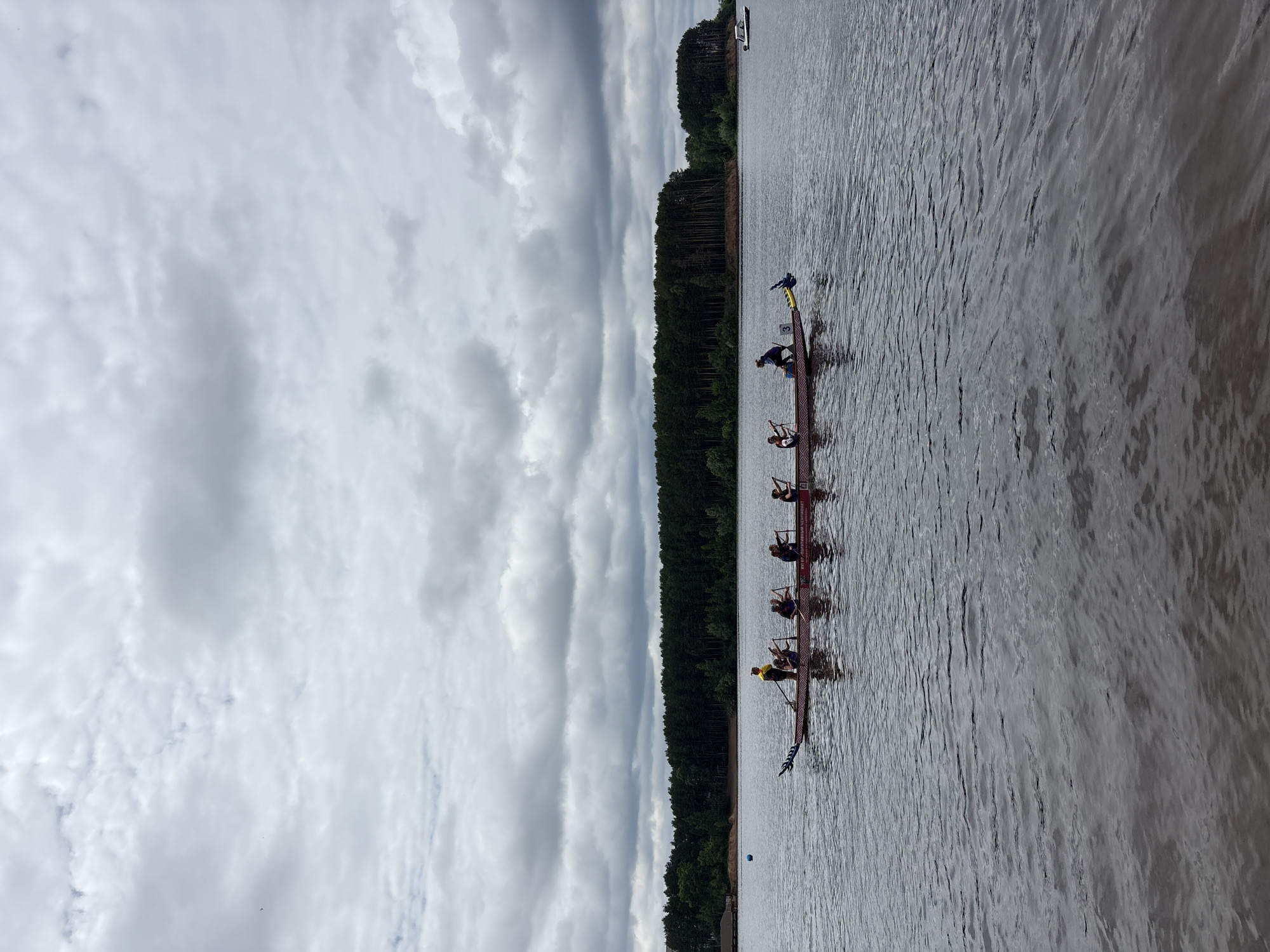 The image depicts a dragon boat race on a body of water, likely the Kama River, in Chaikovsky, Russia. The sky is overcast with thick, gray clouds. In the foreground, the dark water is slightly rippled. A long, red and white dragon boat is positioned horizontally across the frame, with several individuals in it. A person in a yellow shirt stands at the front of the boat, facing backward, appearing to be directing the rowers. Five other individuals are seated in the boat, each holding a paddle and in the process of rowing. The boat has a dragon head and tail decoration. In the mid-ground, a dense forest of tall, dark green trees lines the distant shore. To the far right of the frame, a small white boat is visible on the water. The overall scene suggests a sporting event taking place on a cloudy day.