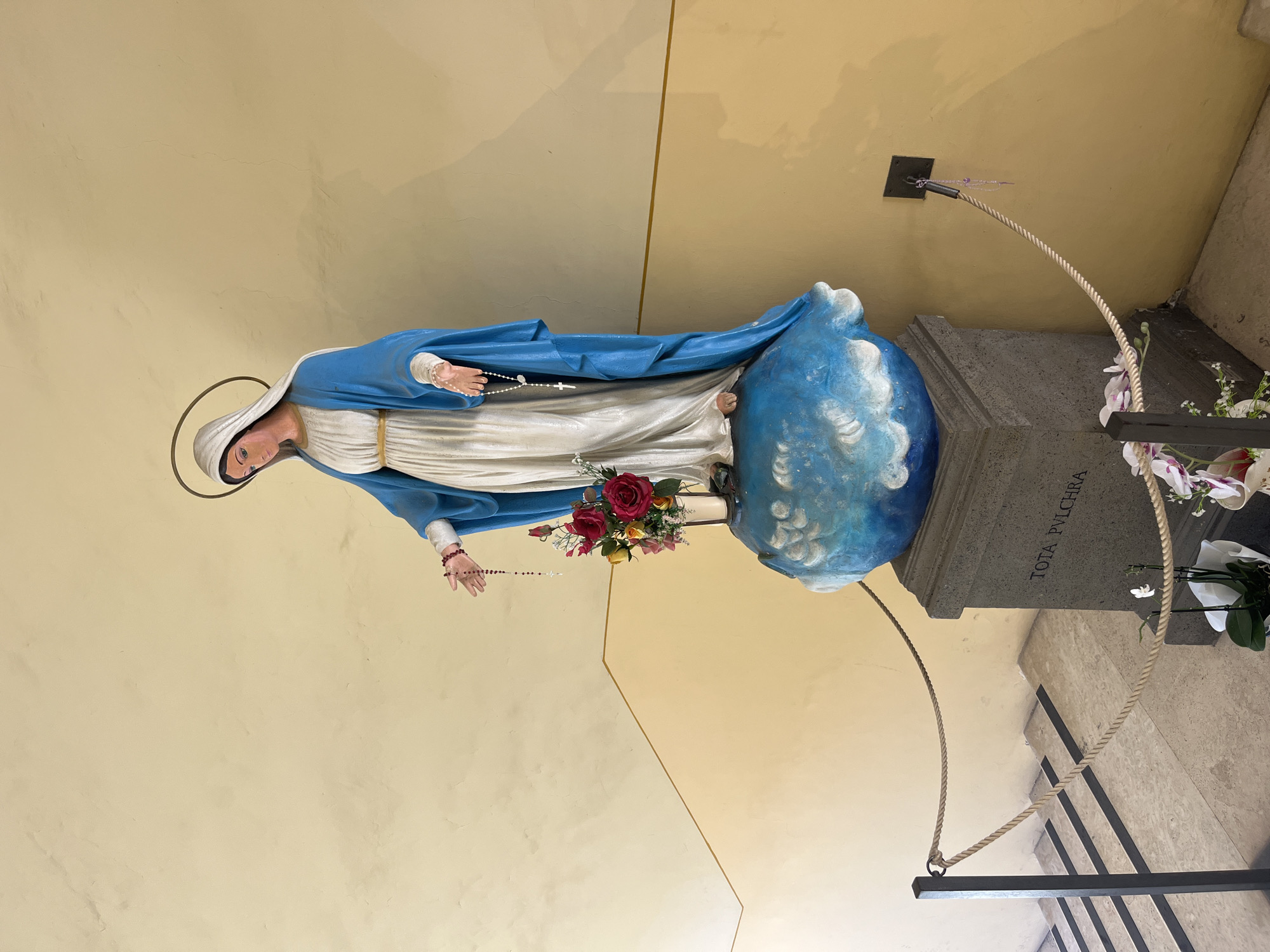 A statue of the Virgin Mary stands on a blue, cloud-like pedestal atop a grey stone base inscribed with "TOTA PVLCHRA." She wears a white dress with a blue mantle and holds a rosary in her right hand. Her left hand is outstretched, holding another rosary with a crucifix. A vase of red and yellow roses sits to her left. The statue is roped off with a light brown rope, supported by black metal posts on the left and a wall anchor on the right. The background is a pale yellow wall with a thin gold line diagonally across it. In the lower left, a staircase with dark stripes on the treads is visible. A small white flower and parts of other flowers are in the foreground. The scene appears to be outdoors or in an open covered area, possibly in a courtyard or passageway.