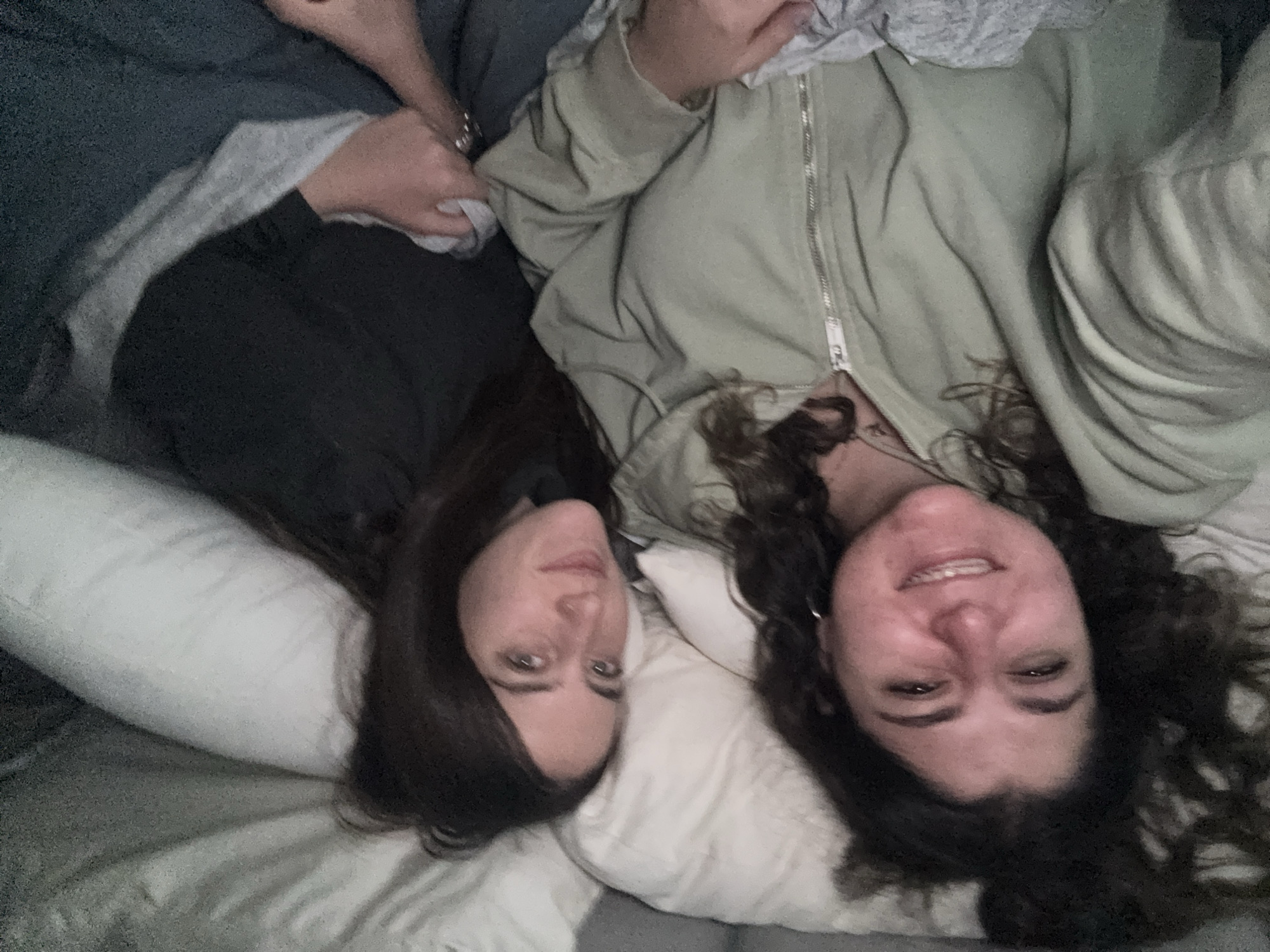 Two women are lying side-by-side on a bed. One woman is on the left side of the frame with dark, straight hair. The other woman is on the right side of the frame with curly, dark hair and wearing a light green zippered jacket. The background shows a white pillow, a white bedsheet, and a gray blanket. The location is undetermined.