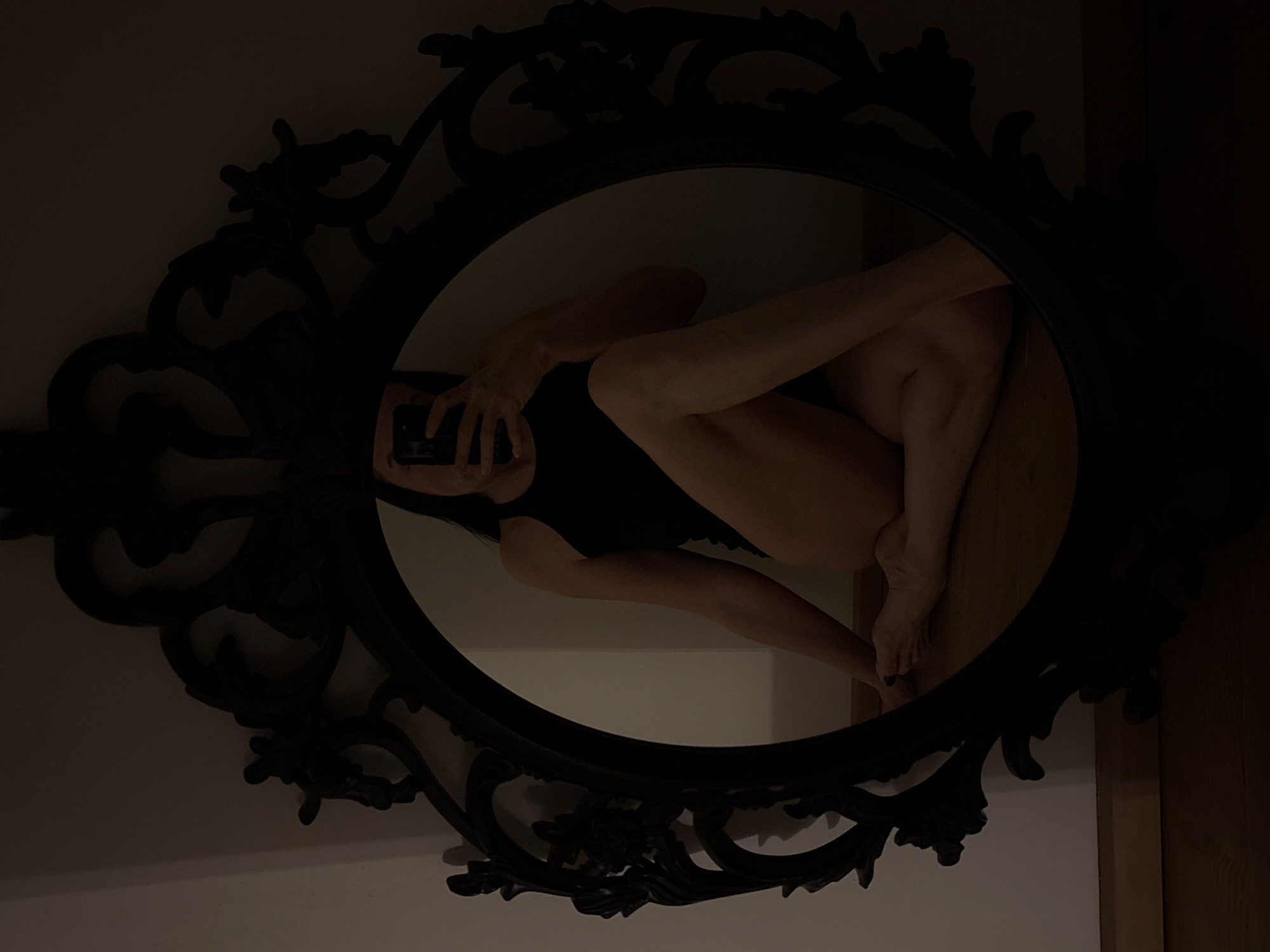 A person in a black garment is visible in a mirror, taking a photograph with a cell phone. The person's legs are bent and drawn up. The oval mirror has a dark, ornate frame. The setting appears to be indoors. The location is unknown and cannot be determined from the image.