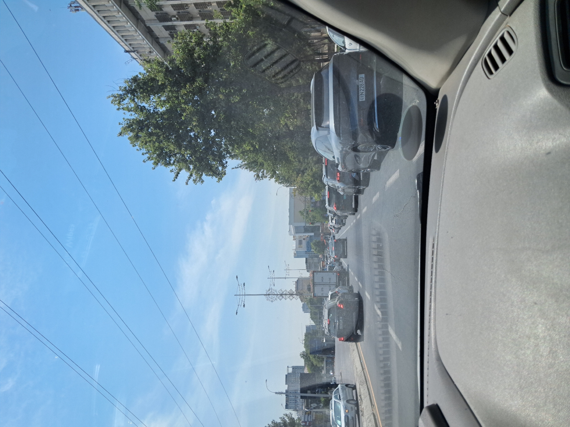 The image captures a traffic jam on a sunny day in Tashkent, Uzbekistan. The perspective is from inside a vehicle, looking forward through the windshield and dashboard. Several cars are visible on a multi-lane road, forming a queue. In the foreground, the dashboard of the car is visible. The middle ground shows the traffic, with vehicles including a black SUV with the license plate "01 N 228 AD", a white car on the left, and a truck with "575 AGA" written on its side. In the background, there are trees, buildings, and overhead power lines against a clear blue sky with scattered clouds. The overall scene depicts a typical urban traffic situation.