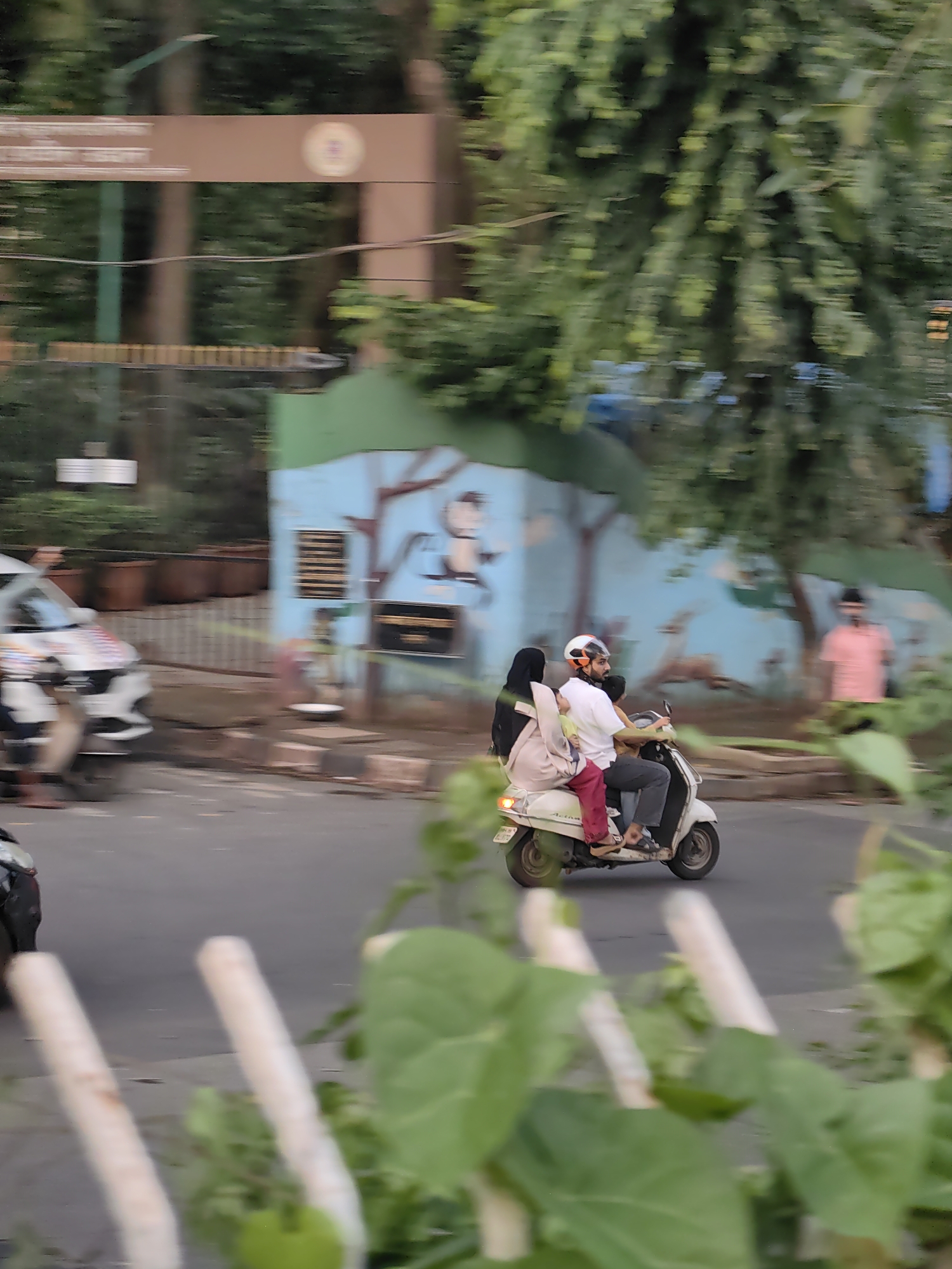 In this image, taken in Mumbai, India, a scooter carrying three people is moving along a street. The driver, a man wearing a white t-shirt and a helmet, is at the front. Behind him, a woman in a light-colored hijab and a black garment is seated, holding a young child. The child appears to be wearing a pink outfit. The scooter is white and appears to be a Honda Activa.
The street is paved, and there are planters with some vegetation visible behind a metal railing on the left side of the road. Further to the left, a white car with red and blue stripes is partially visible.
In the background, a light blue wall with a mural is prominent. The mural depicts stylized trees and animals, possibly a deer or antelope. A man in a pink shirt stands near the mural. Above the mural and to the left, a brown sign with text and a circular emblem is mounted on a structure. Trees with green foliage are visible behind and above the mural.
The foreground is blurred, showing parts of large green leaves and light-colored vertical posts, suggesting the photo was taken from behind a fence or foliage. The overall impression is of a street scene with movement and a blend of urban elements and natural growth. The image has a slight motion blur, indicating that the photograph was taken while the camera or the subjects were in motion.