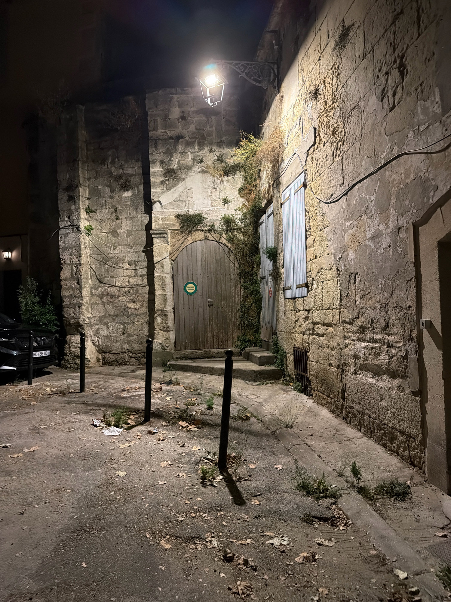 The image depicts a narrow street at night in Arles, France. A vintage streetlamp illuminates a cobblestone pathway and the rough-hewn stone walls of buildings. A large wooden double door, possibly an entrance to a courtyard or building, is visible on the left. The door has a green circular sign with text in French. To the right of the door, a set of wooden shutters covers a window. A black car is parked on the left side of the street. The ground is littered with fallen leaves and debris. The scene suggests an old part of the city, possibly a historic district.