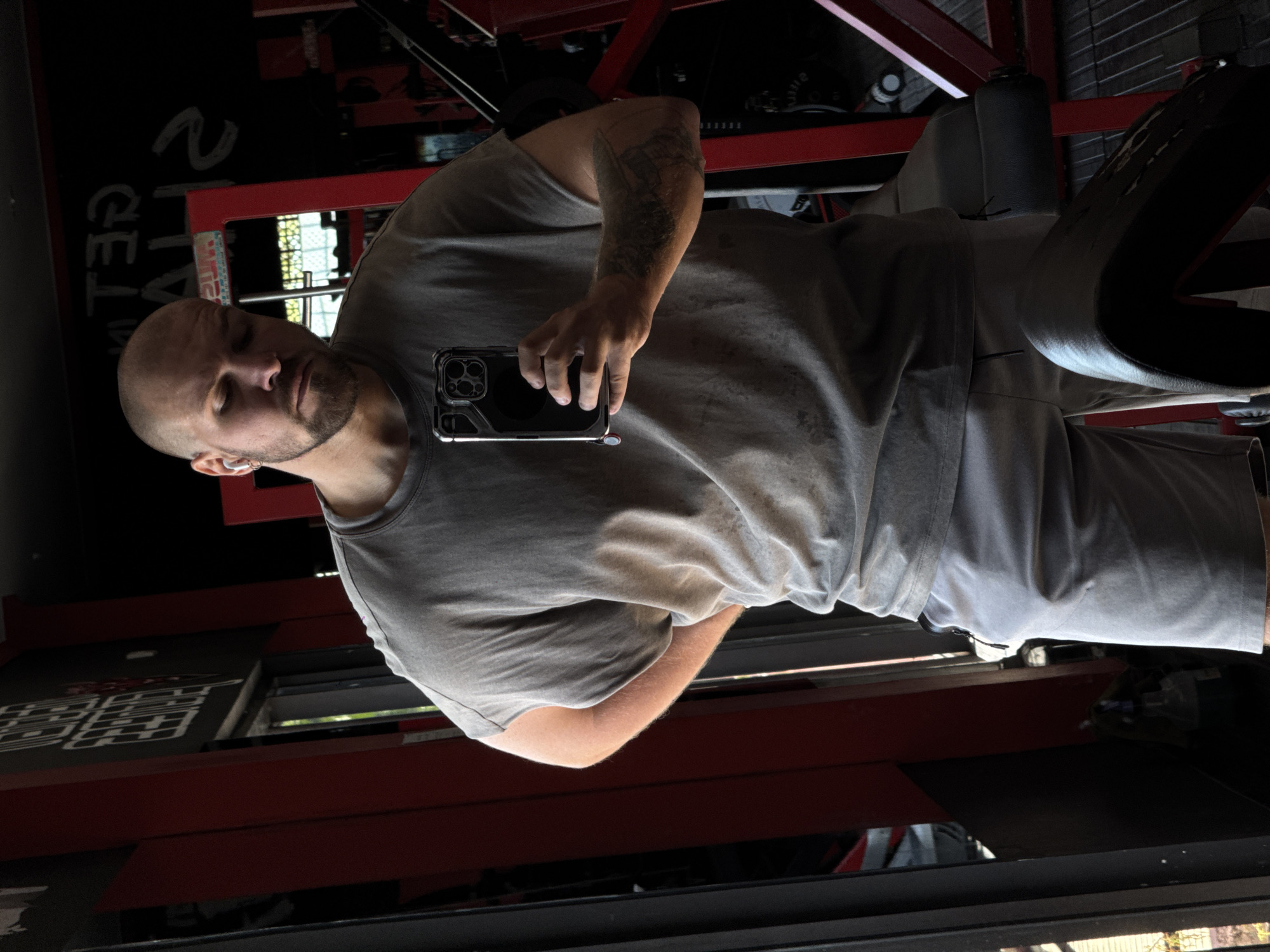 A bald, Caucasian man with a beard, wearing a grey t-shirt and grey shorts, is standing in a gym, taking a mirror selfie. He has white wireless earbuds in his ears and a tattoo on his left arm. The gym appears to be well-equipped, with red metal weight machines visible in the background. A window with greenery outside is visible to the left of the man, suggesting a location that receives natural light. The overall atmosphere is casual and focused on fitness. The man's posture and expression convey a sense of confidence and self-awareness. The lighting is somewhat dim, with shadows cast by the equipment.